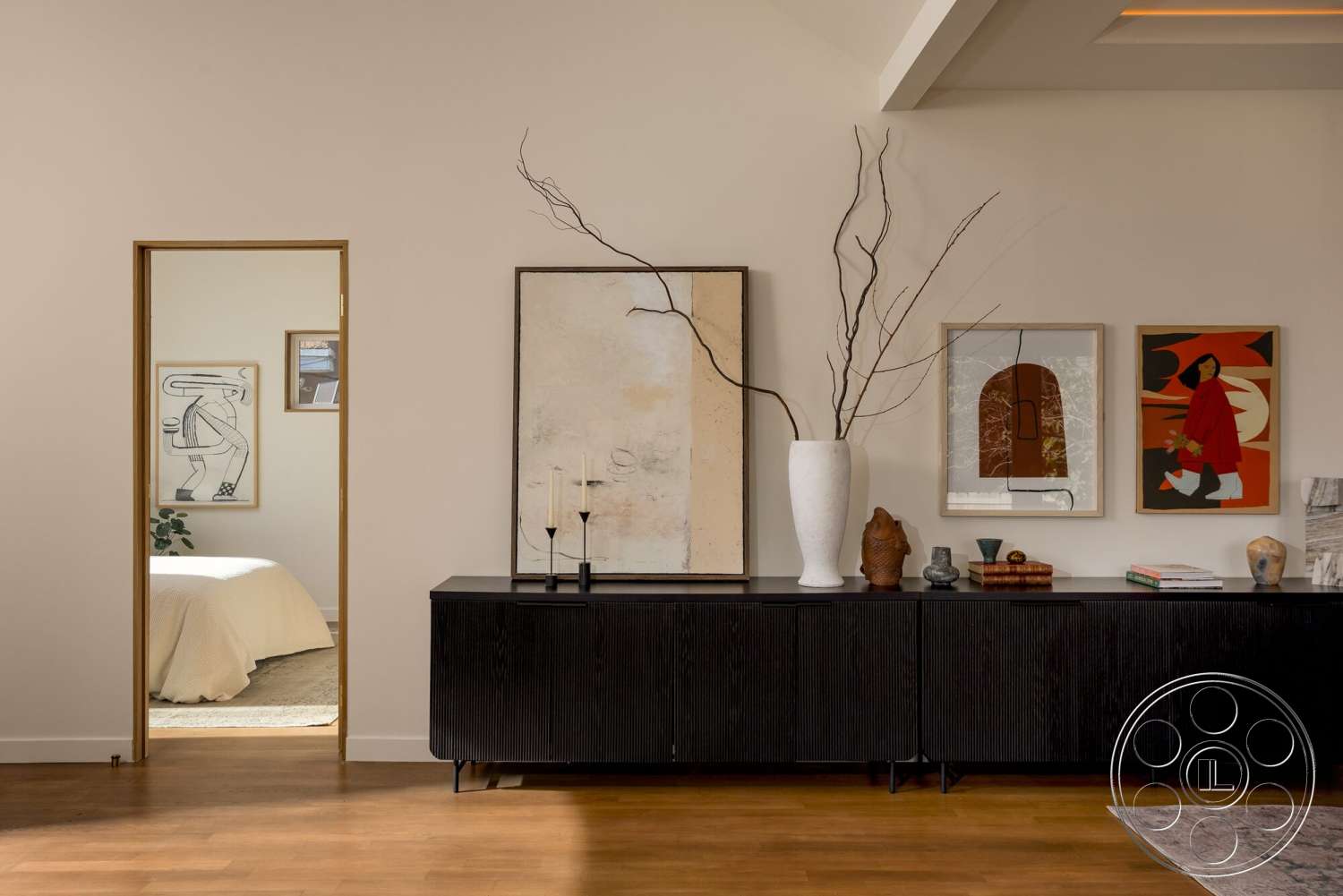Craftsman 65 - textured plaster walls, open doorway, natural light from window, hardwood living room floors, framed abstract wall art, modern sideboard with slatted design, large white ceramic vase, minimalist decorative accessories, light brown bedding visible in adjacent room