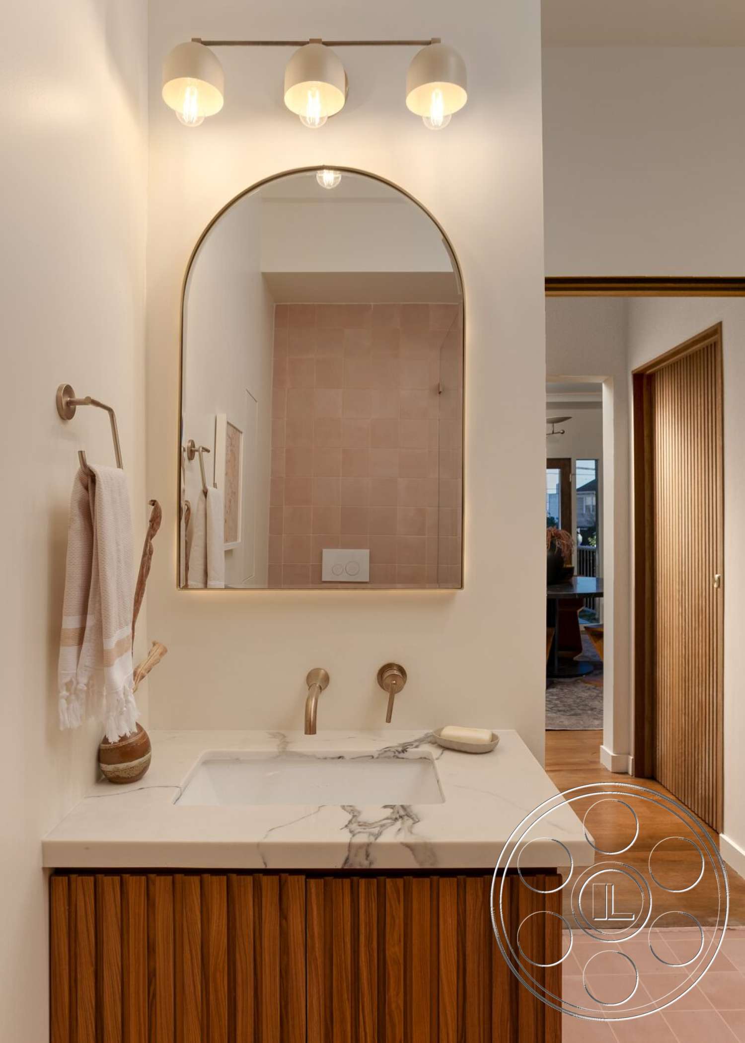 Craftsman 65 - wood cabinetry, modern light fixtures, concrete flooring, marble countertop, textured wall treatment, dark wood door, arched mirror, white painted walls, brass faucet fixtures, pink tile backsplash