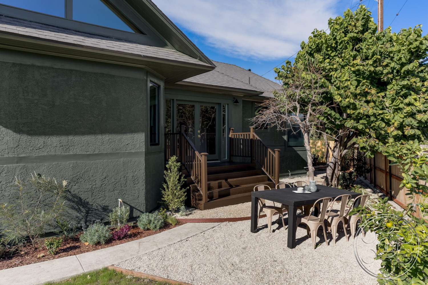 Craftsman 65 - exterior, large glass windows, green foliage, single-story home, gabled roof, textured plaster walls, rectangular dining table, metal dining chairs, landscaped garden beds, urban backyard setting, wooden deck stairs, gray gravel patio flooring