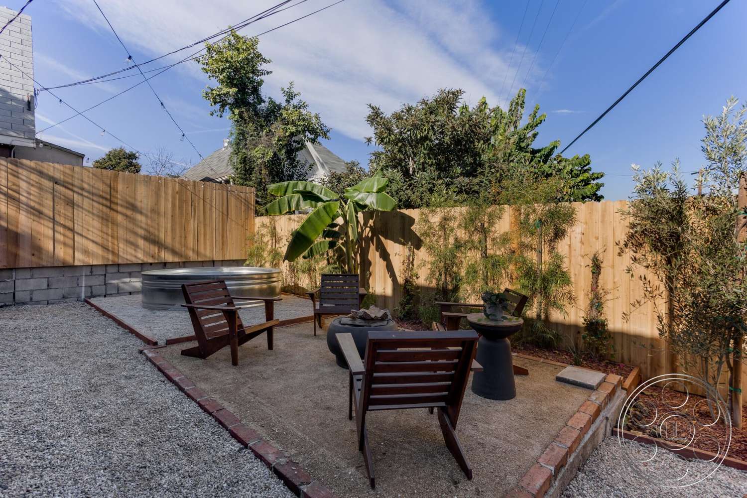 Craftsman 65 - exterior, concrete block walls, wooden privacy fence, wooden Adirondack chairs, gravel patio flooring, landscaped garden beds, lush greenery surrounding, string lights overhead, metal fire pit, rectangular sitting area, brick border landscape, potted banana plant
