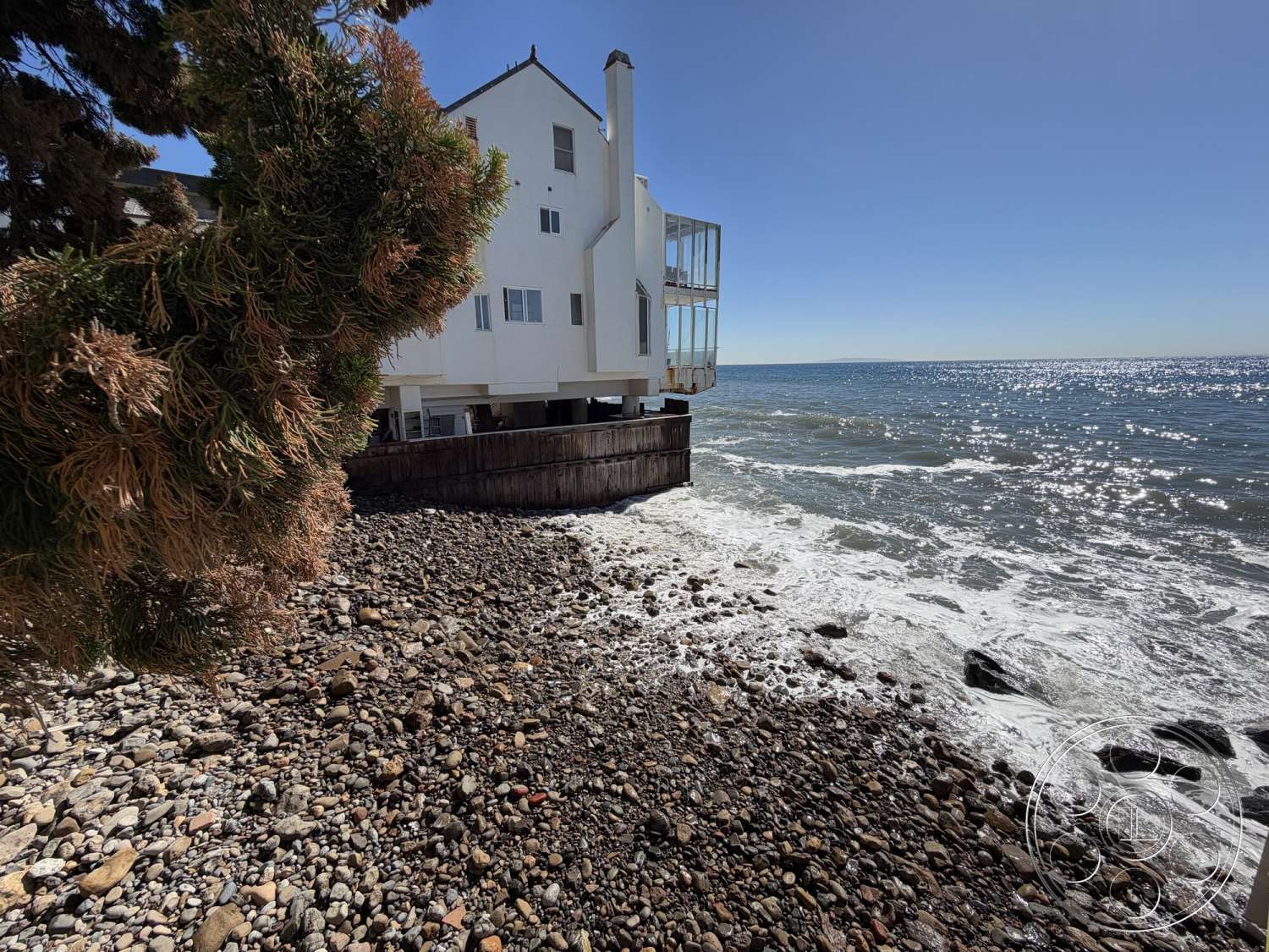 The Rittshouse - exterior, white stucco walls, large glass windows, rocky shoreline, panoramic ocean view, surrounding natural landscape, angular roof shape, wooden coastal decking, elevated house structure, two-story design