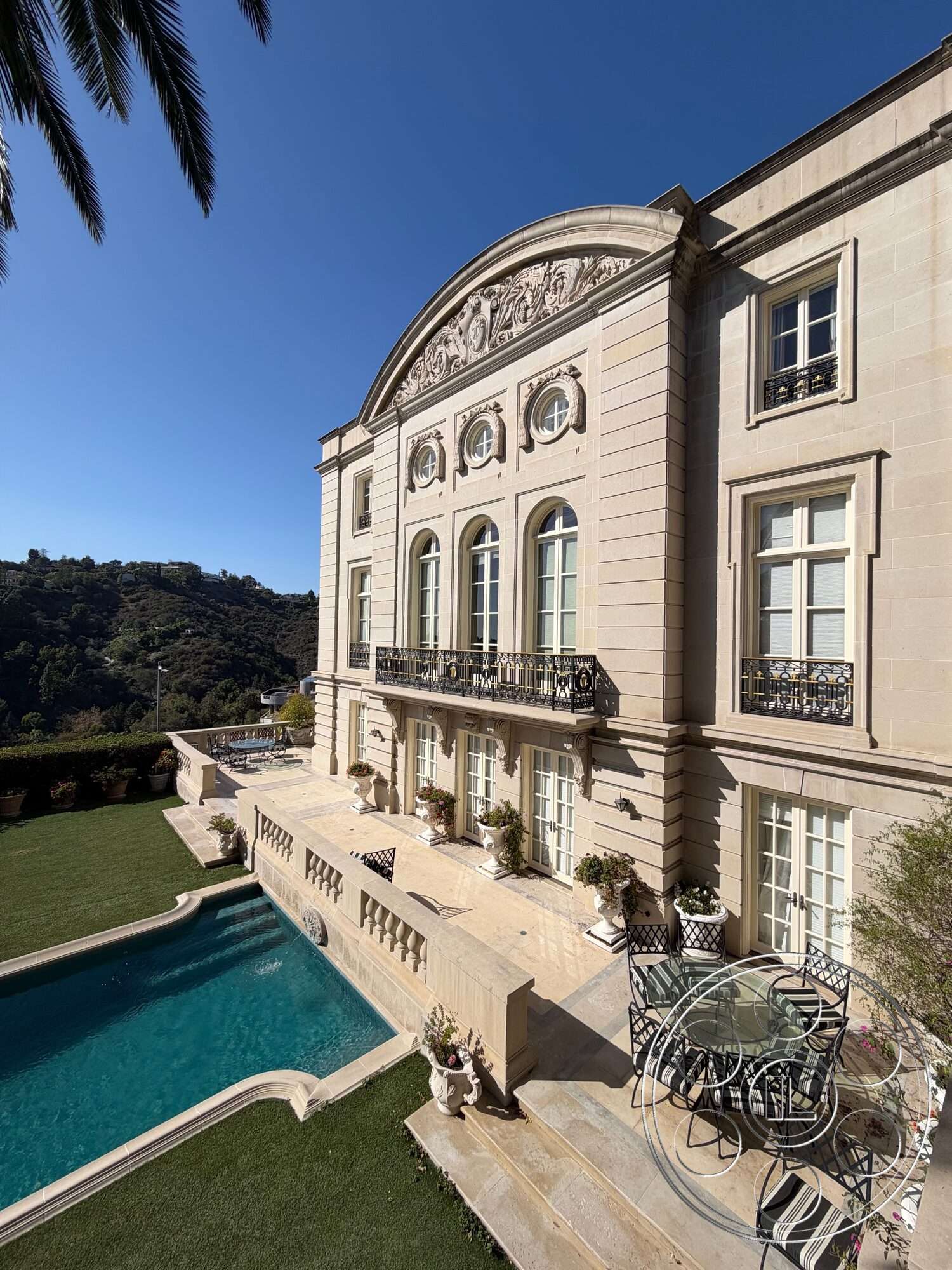 Mansion 139 - exterior, rectangular swimming pool, manicured lawn, outdoor dining set, modern Mediterranean architecture, landscaped garden area, sunny outdoor environment, arched window details, marble patio flooring, ornate balcony railings, decorative stone cornice, limestone façade, double-hung windows with shutters, potted plants on terrace, French-style doors, views of hillside