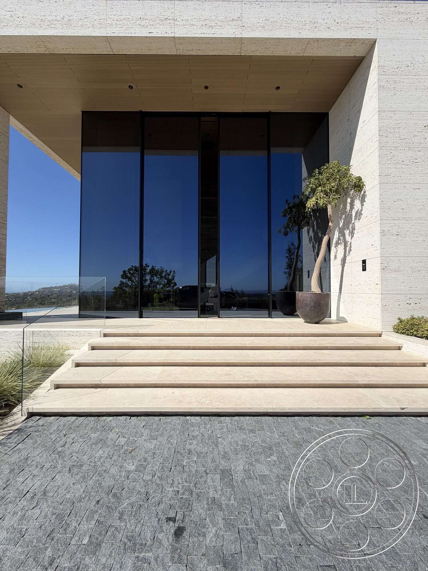 CK Mansion - exterior, modern architectural style, floor-to-ceiling glass windows, wood paneled ceiling, contemporary entrance, limestone steps, linear planters, glass front doors, textured stone paver driveway, landscape with minimal greenery