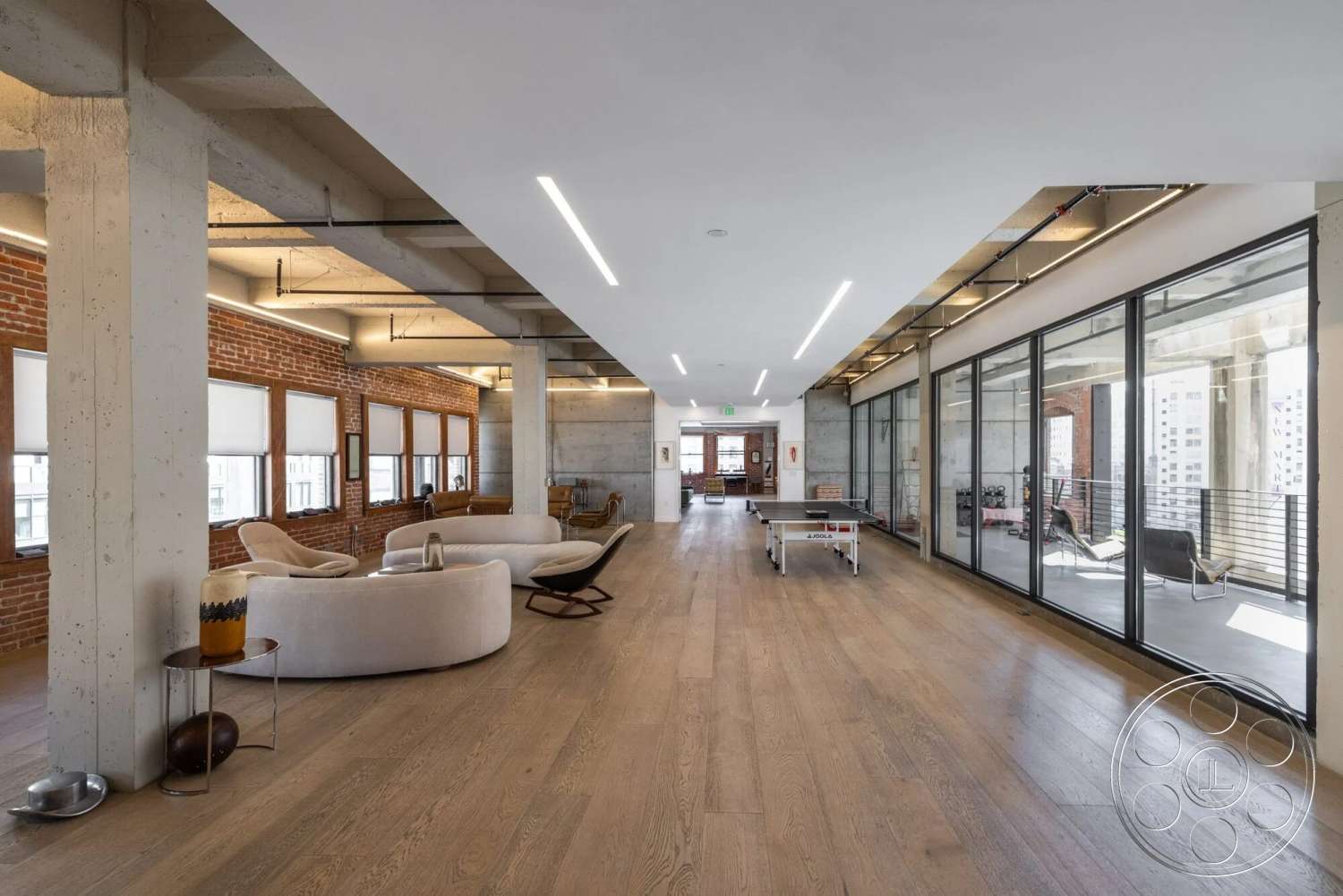 Loft 1 - hardwood flooring, open floor plan, minimalist furniture, functional workspace, contemporary design elements, modern lounge area, ping pong table, glass wall partitions, exposed brick accent wall, industrial style interior, large windows with city view, linear LED ceiling lights, urban loft setting, concrete structural columns, living room with seating area