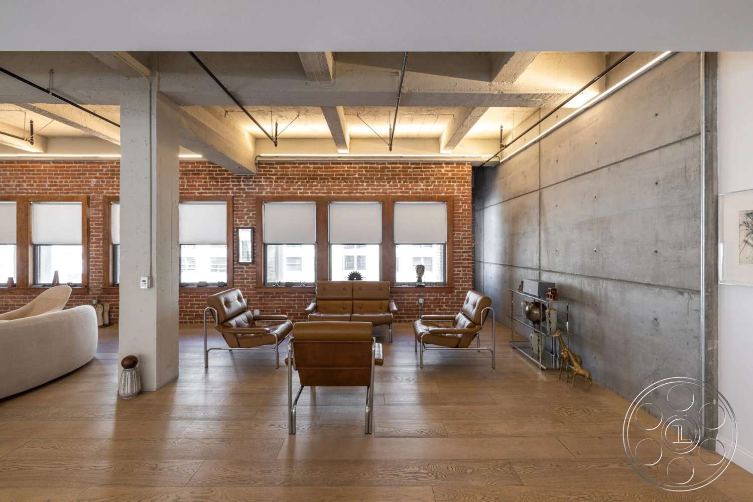 Loft 1 - open floor plan, concrete walls, concrete ceiling, metal side table, hardwood living room floors, exposed brick accent wall, industrial-style lighting, leather lounge chairs, large windows with roller shades