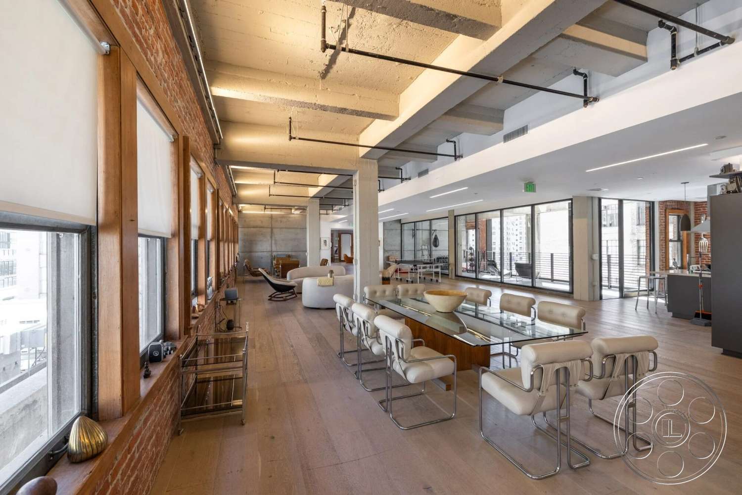 Loft 1 - natural light, urban setting, floor-to-ceiling windows, glass dining table, concrete ceiling, modern chrome dining chairs, open floor plan interior, exposed brick accent wall, industrial-style lighting fixtures, light wood plank flooring, rectangular dining area, modern kitchen area, steel brackets and piping, contemporary loft style