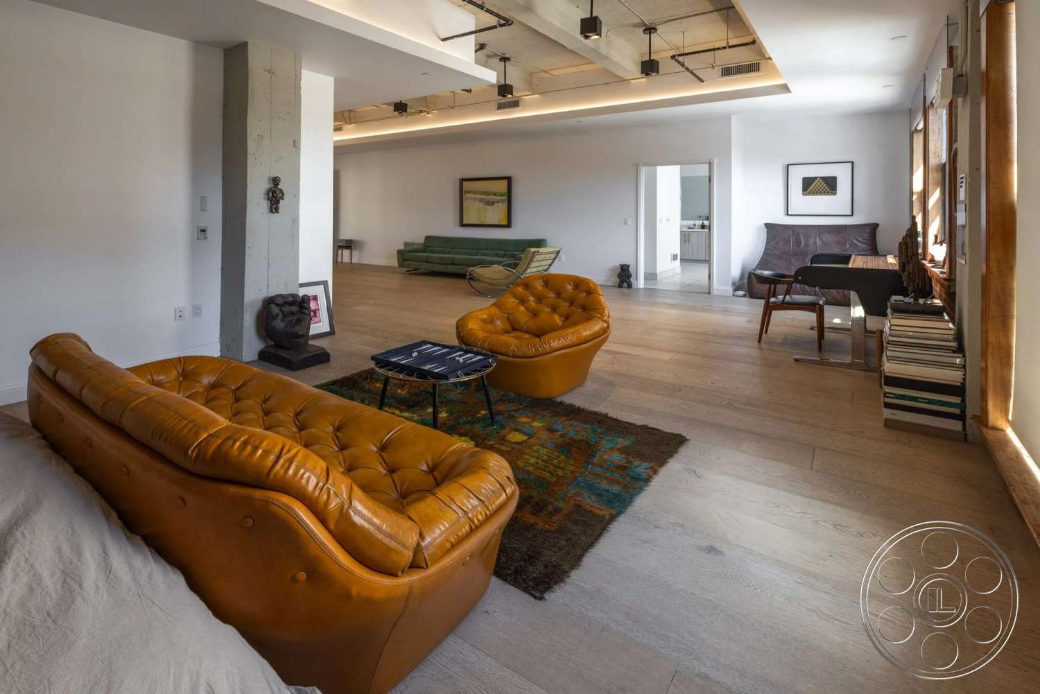 Loft 1 - textured area rug, open floor plan, modern art wall decor, natural light from large windows, exposed ceiling beams, dark wood shelving, contemporary coffee table, hardwood living room floors, concrete accent wall, neutral white painted walls, tan leather tufted sofas, green velvet couch, minimalist study area