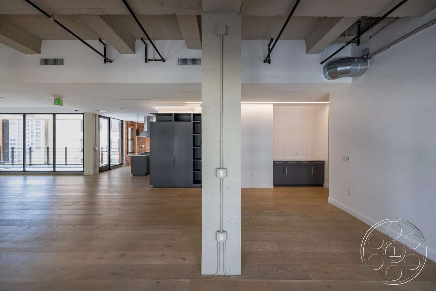 Loft 1 - light-colored walls, open floor plan, brick accent wall, large floor-to-ceiling windows, metal ductwork, modern kitchen design, wooden plank flooring, exposed concrete pillars, industrial ceiling details