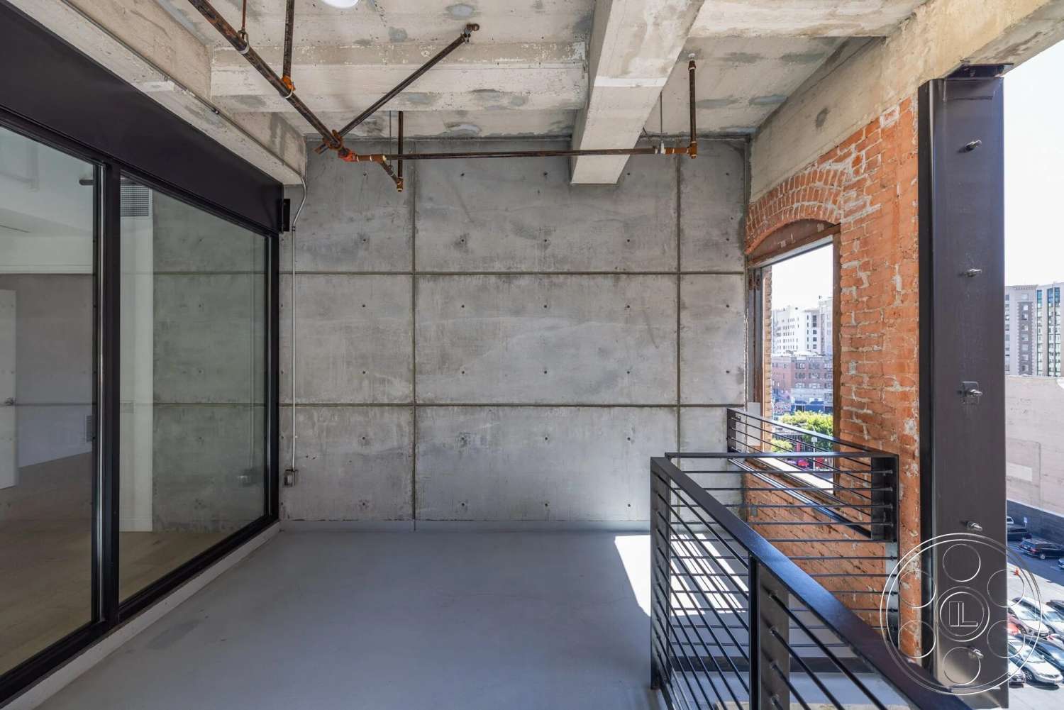Loft 1 - large glass sliding doors, open floor plan, concrete flooring, exposed concrete walls, city skyline view, steel support beams, modern industrial style, industrial balcony railing, red exposed brick accent wall, interior balcony space