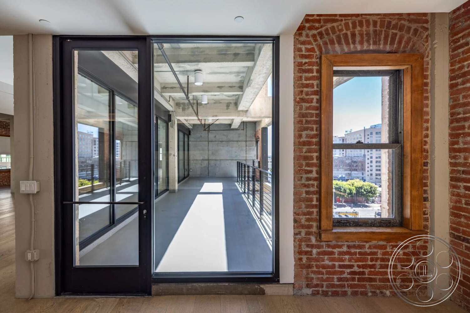 Loft 1 - modern architectural style, exposed concrete ceiling, large glass door, natural light from floor-to-ceiling windows, urban setting view, exterior windows with black metal frames, brick wall with red brick finish, hardwood floor in light finish, concrete flooring in open area, vertical railing, wood trim on window