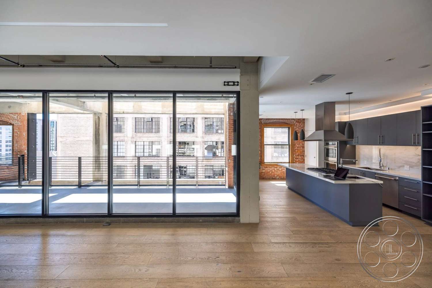 Loft 1 - dark wood cabinetry, glass sliding doors, pendant lighting fixtures, stainless steel kitchen appliances, quartz kitchen countertops, open floor plan interior, exterior balcony, exposed brick accent wall, large windows with city view, industrial style ceiling, urban loft setting, hardwood floor living area, modern kitchen with island, steel balcony railing, concrete structural columns