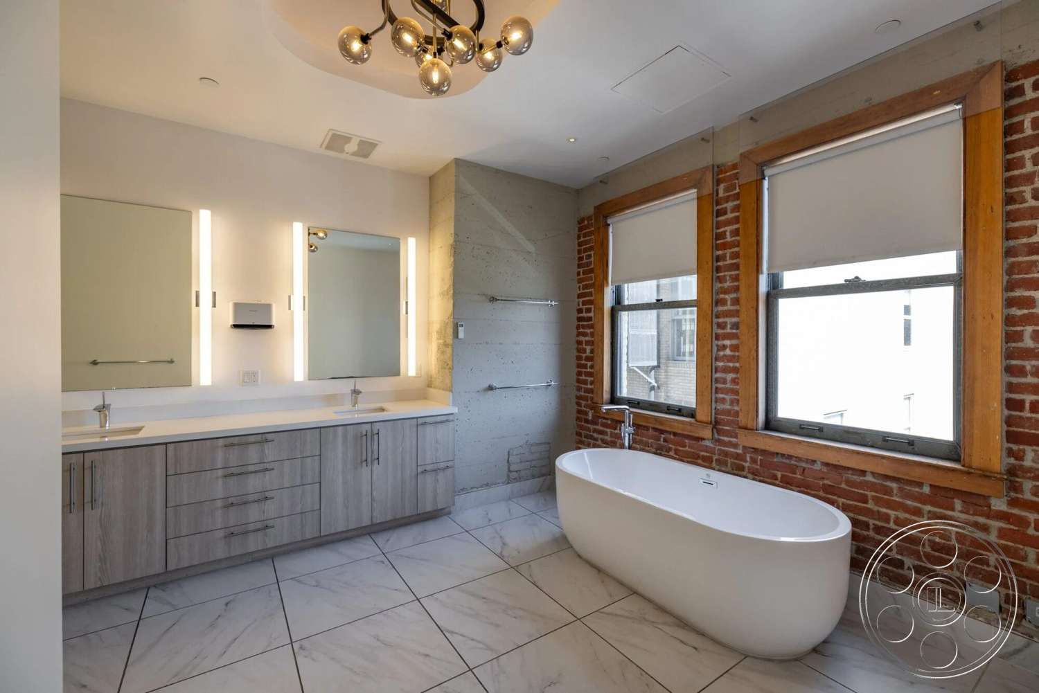 Loft 1 - metal fixtures, textured plaster ceiling, white freestanding bathtub, interior bathroom, exposed red brick accent wall, white marble floor tiles, concrete shower wall, light wood vanity cabinets, dual bathroom sinks, round chandelier lighting, glass mirror with built-in lights