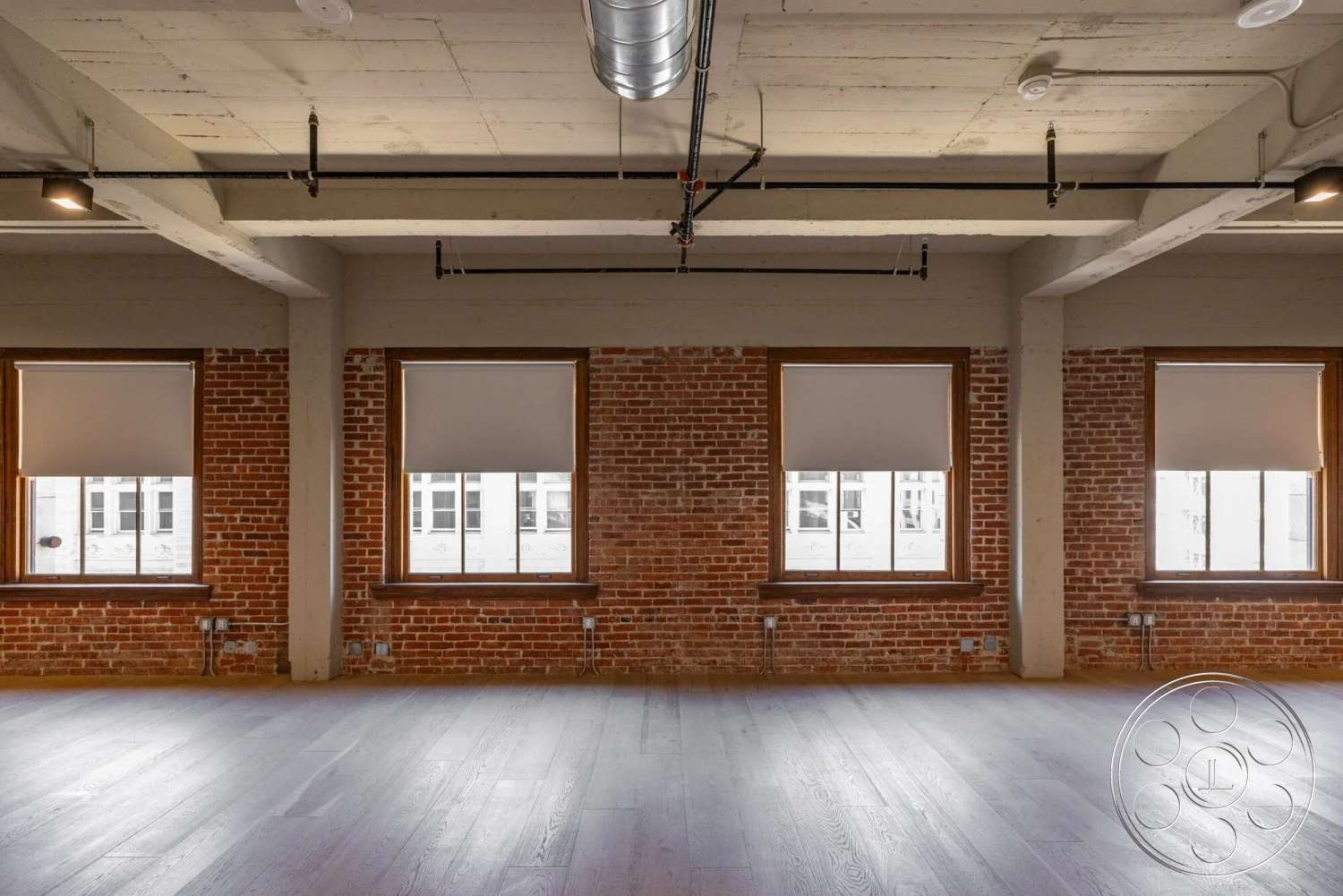 Loft 1 - natural light, hardwood flooring, open floor plan, track lighting, exposed ductwork, concrete ceiling, wood window frames, modern minimalist aesthetic, industrial style interior, urban loft setting, exposed brick accent walls, large windows with wood trim
