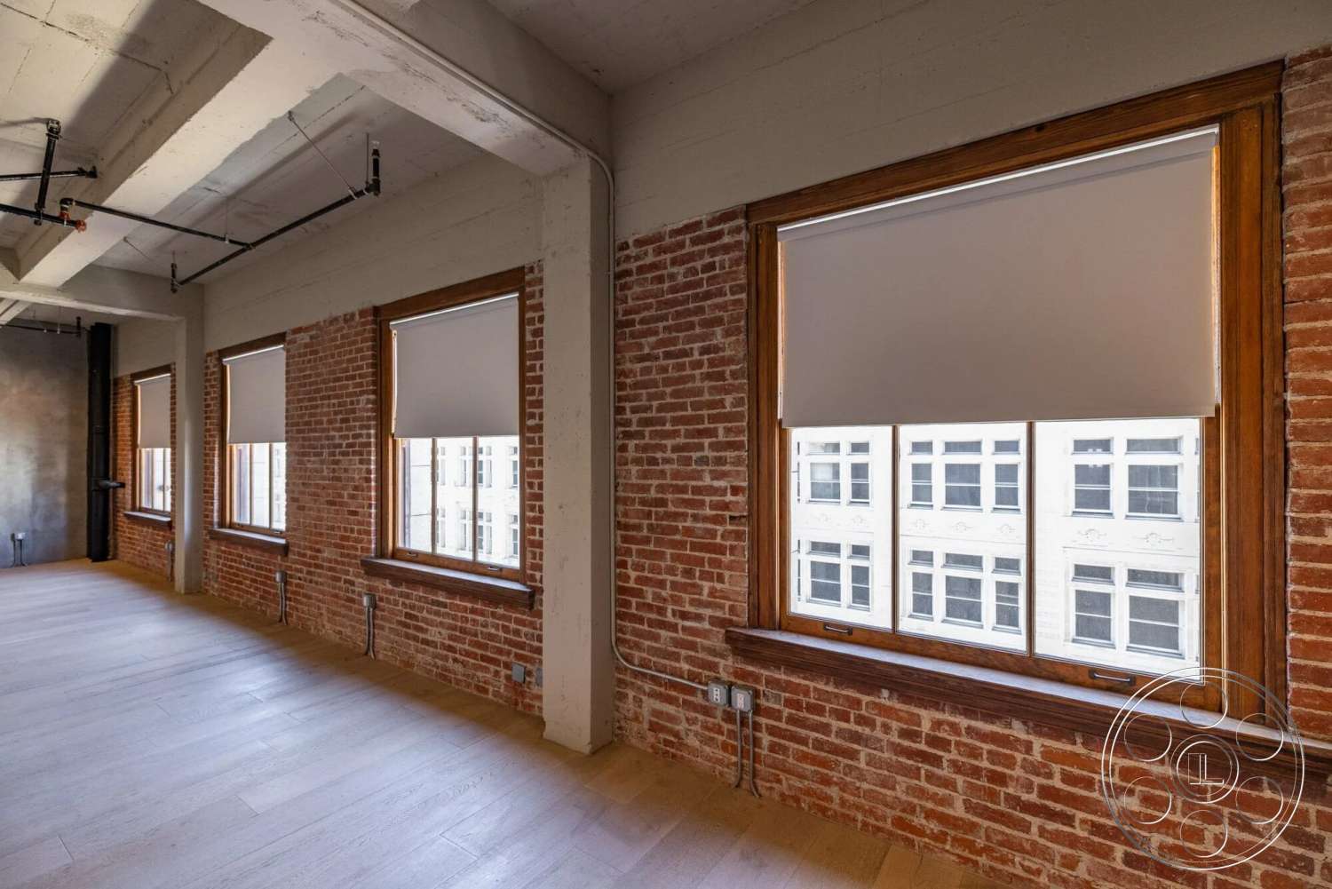 Loft 1 - neutral color palette, hardwood flooring, open floor plan, studio space, urban loft, concrete ceiling, exposed piping, wood window frames, exposed brick accent wall, natural light ambiance, minimalist interior, industrial style windows, white roller shades