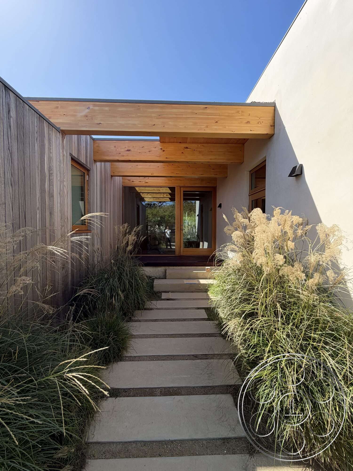 Retro House - exterior, modern architectural style, textured plaster walls, horizontal wood siding, wood beam pergola, large glass entrance doors, minimalist landscape design, natural light entrance, concrete pathway with gravel borders, landscaped grass borders