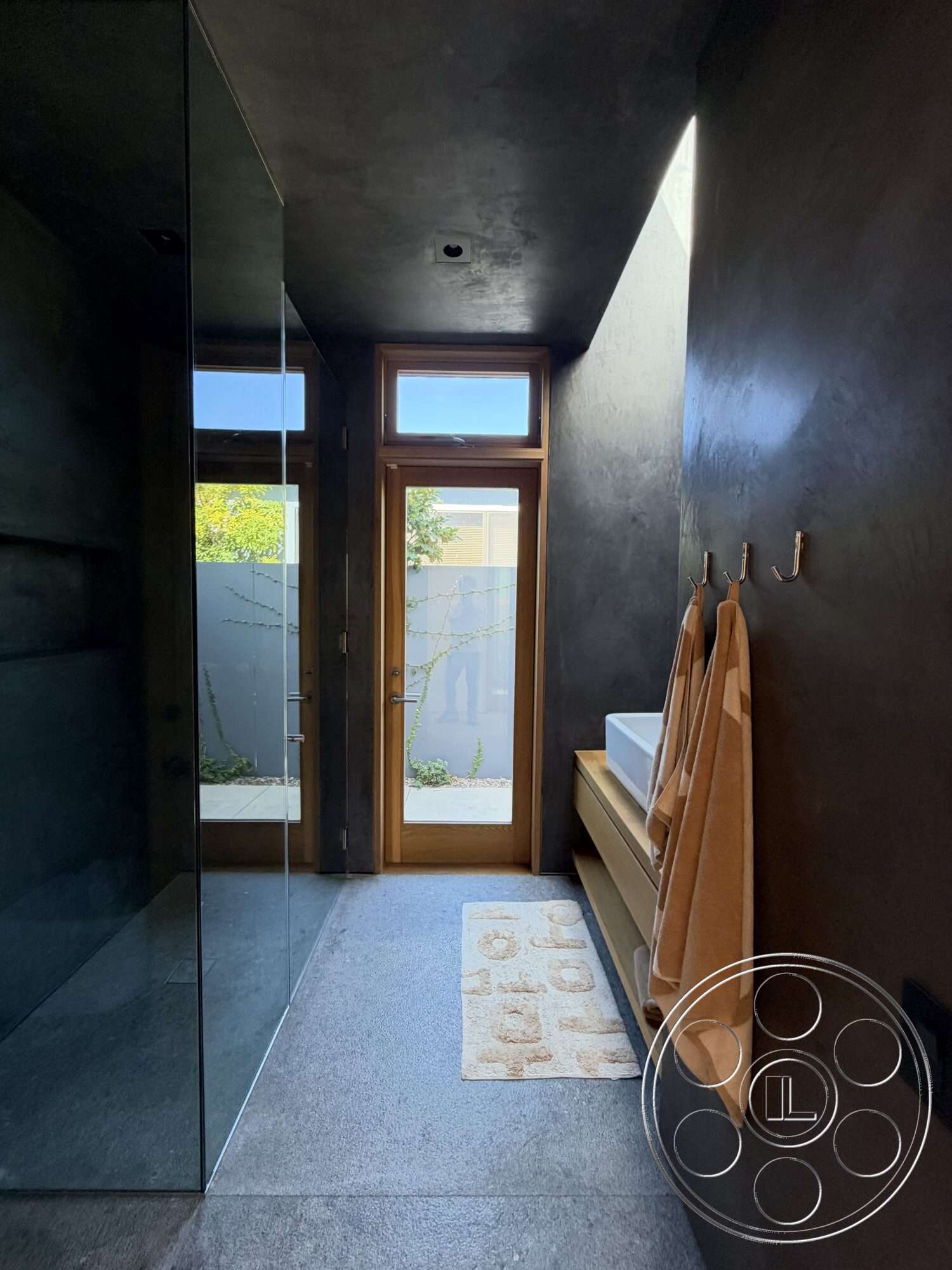 Retro House - glass shower enclosure, minimalist design elements, textured concrete walls, modern architectural style, natural light from windows, towel hooks, wooden door frame, concrete bathroom flooring, floating wood vanity, white sink basin, plush bathroom mat