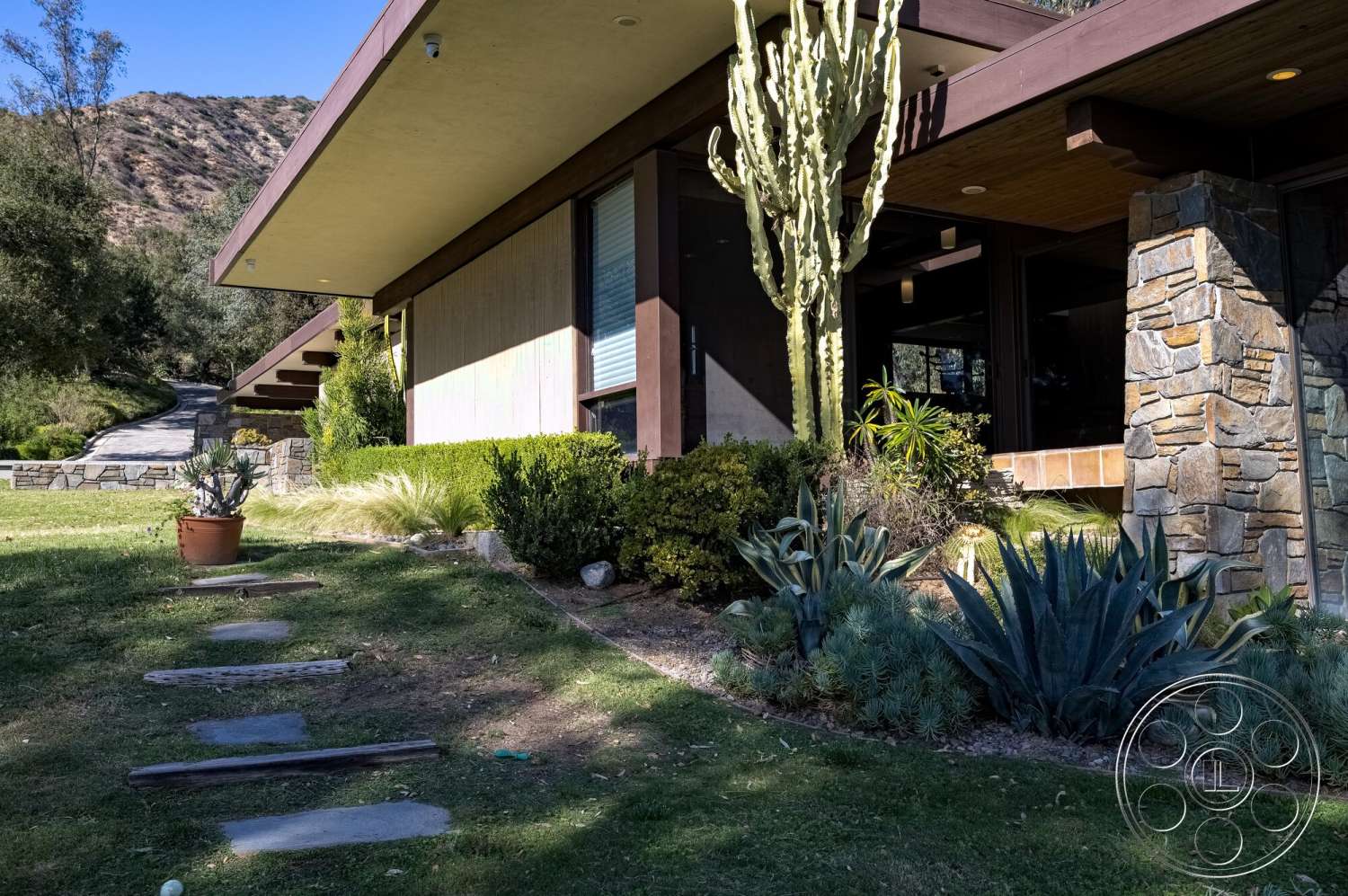 MCM 29 - exterior, flat roof, lush greenery, large glass windows, concrete pathway, sloped driveway, desert landscape, cactus plants, stone feature wall, planted garden beds, decorative stone planters, mid-century modern architecture, wood beam overhang, natural stone landscaping, deck with wood railing