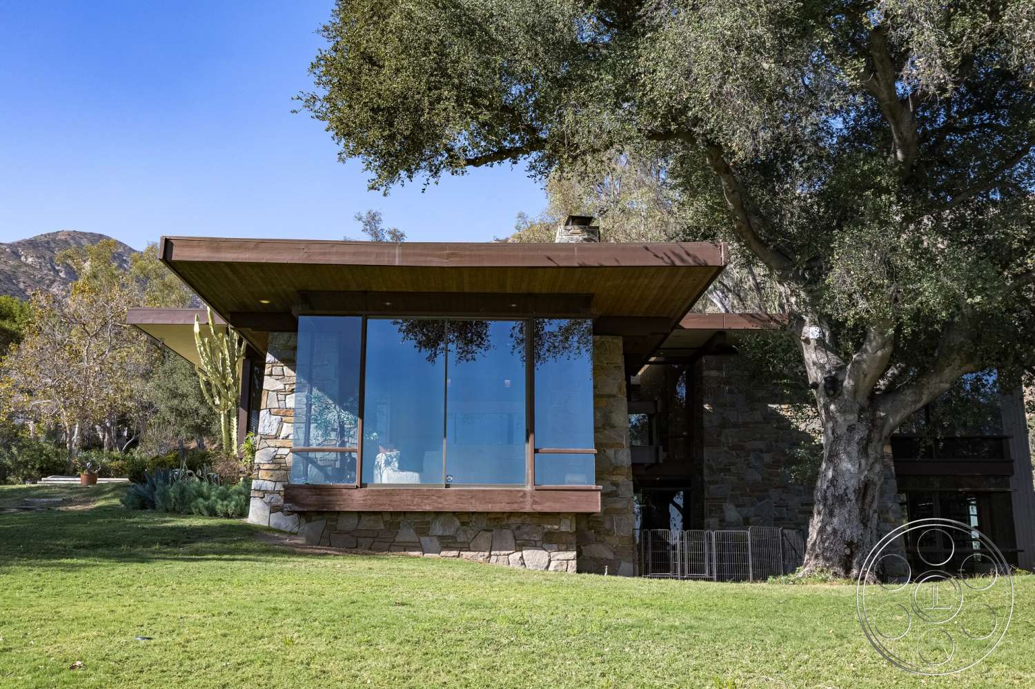 MCM 29 - exterior, modern architectural style, mountain backdrop, large floor-to-ceiling windows, flat roof design, wood beam roof overhang, stone exterior walls, green lawn setting, landscape with shrubs and trees