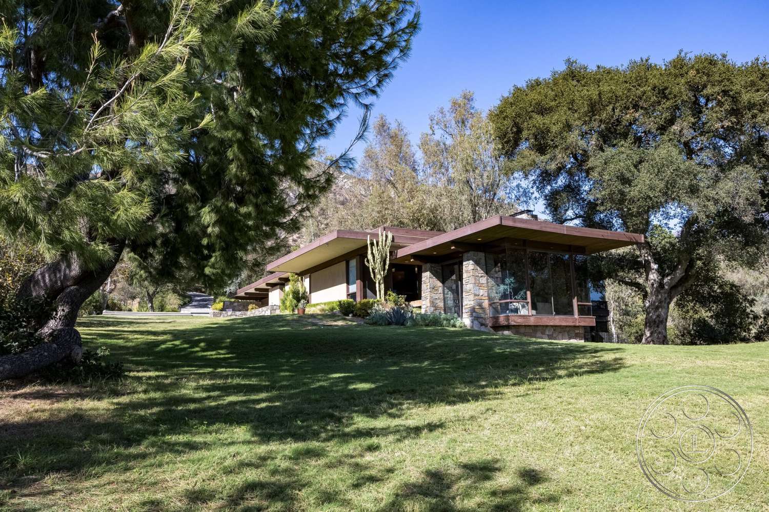 MCM 29 - exterior, large glass windows, surrounding trees, landscaped front yard, lawn area, stone feature wall, mid-century modern architecture, wood roof overhang, sloped grass landscape
