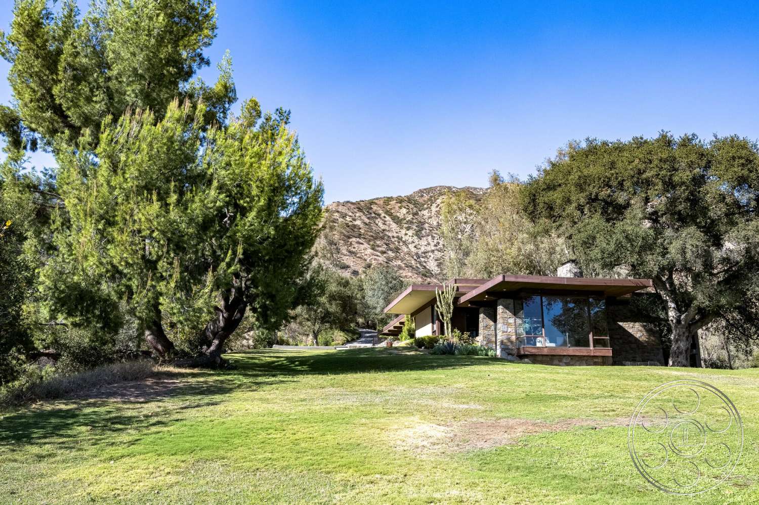 MCM 29 - exterior, large glass windows, modern architectural style, open outdoor space, mountain backdrop, natural setting, stone facade, flat roof design, landscaped lawn area, horizontal wood overhang