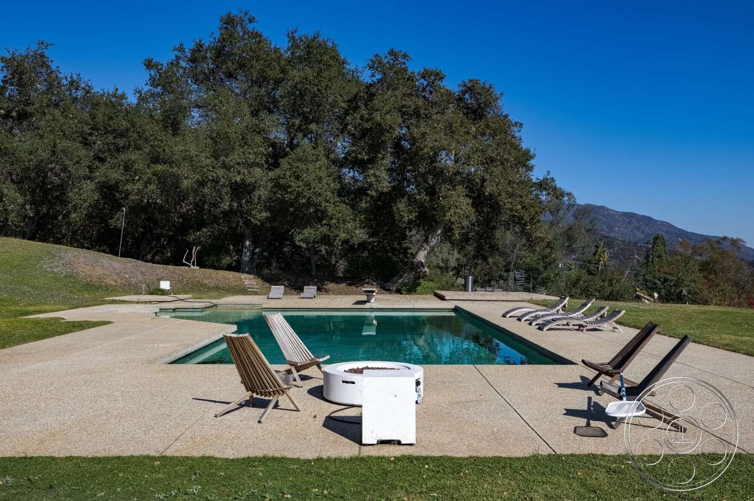MCM 29 - exterior, surrounding greenery, outdoor living space, lawn area, poolside lounge chairs, rectangular pool, concrete pool border, sunny outdoor environment, mountain view backdrop, exposed aggregate concrete patio flooring, shallow end of pool