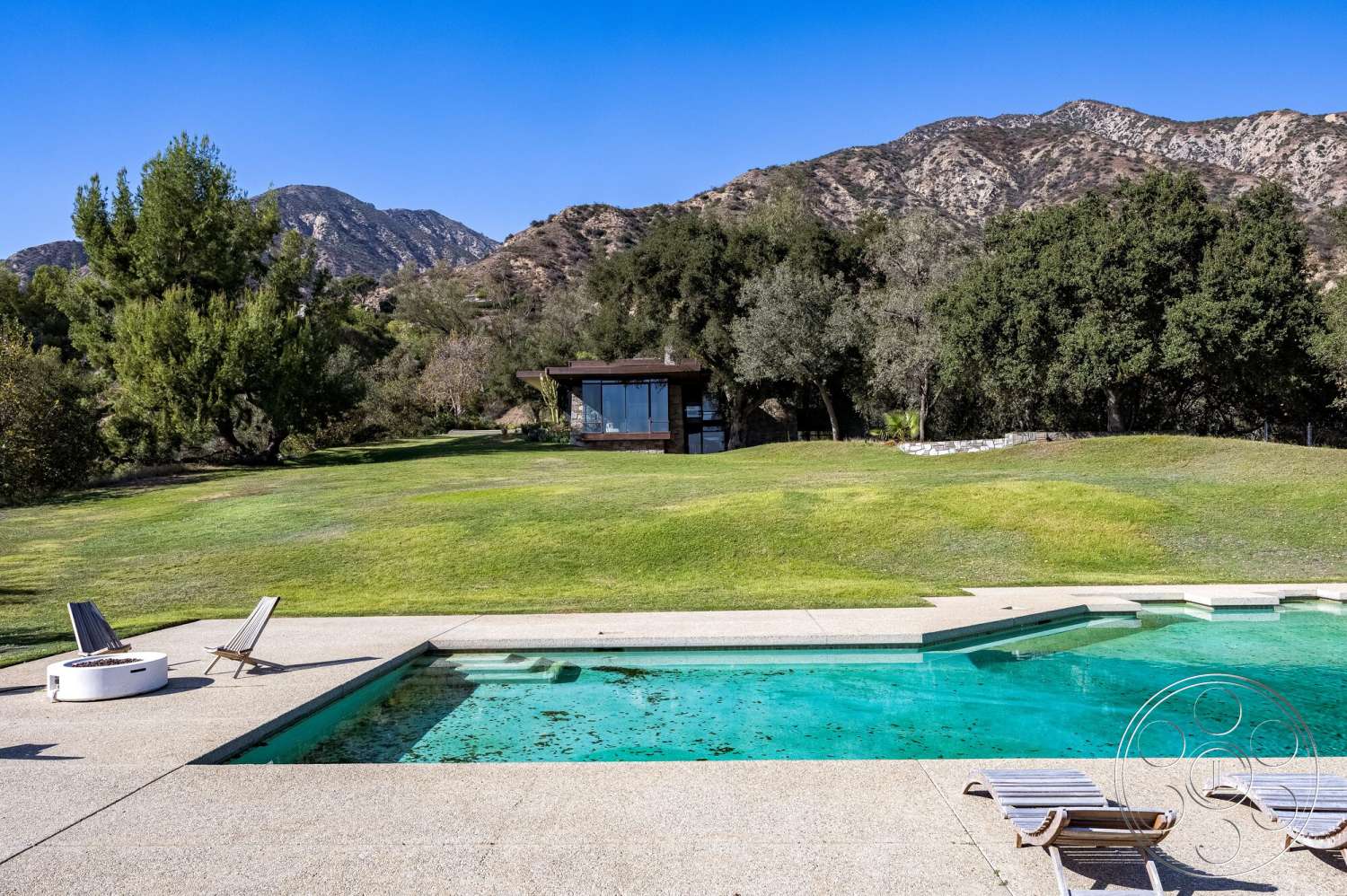 MCM 29 - exterior, landscaped garden, grass lawn, mountain view, outdoor lounging area, concrete pool deck, rectangular pool, wooden lounge chairs, privacy trees, natural stone border, open landscape setting, modern glass house