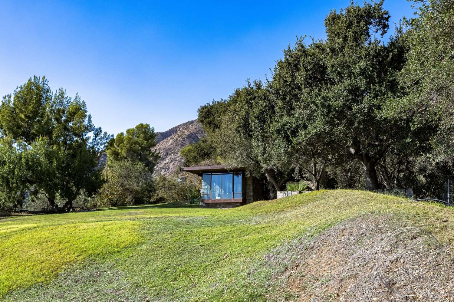 MCM 29 - exterior, outdoor space, suburban setting, landscaped lawn, tall trees, mountain backdrop, modern cabin, open sky, glass panel walls, natural foliage, wood framing, rolling hill terrain