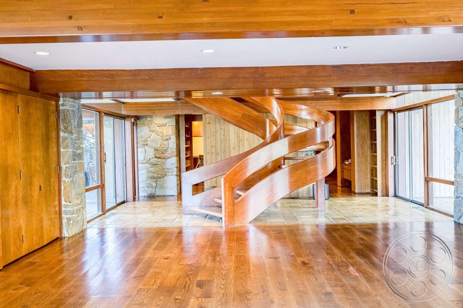 MCM 29 - stone accent wall, hardwood floor, glass door access, exposed wood ceiling beams, natural light through large windows, textured wood paneling, spiral wooden staircase