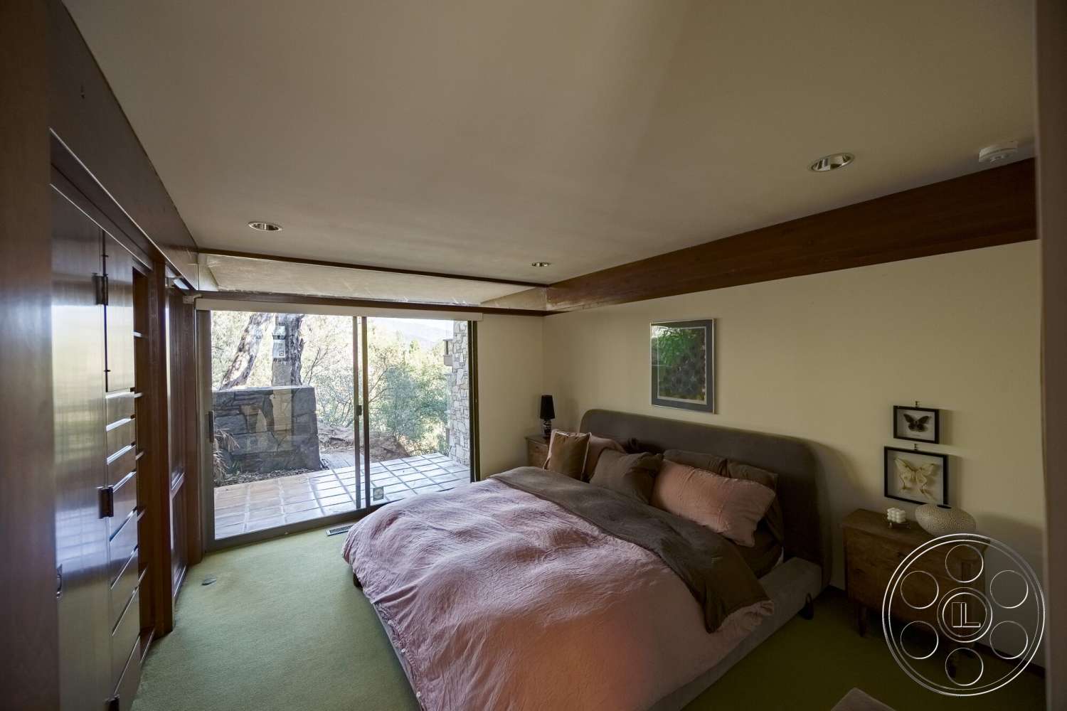 MCM 29 - decorative wall art, textured plaster walls, glass sliding door, wood beam ceiling, green carpet bedroom flooring, wood paneling built-in wardrobe, stone patio view, queen-size bed with plush bedding, simple wooden bedside table