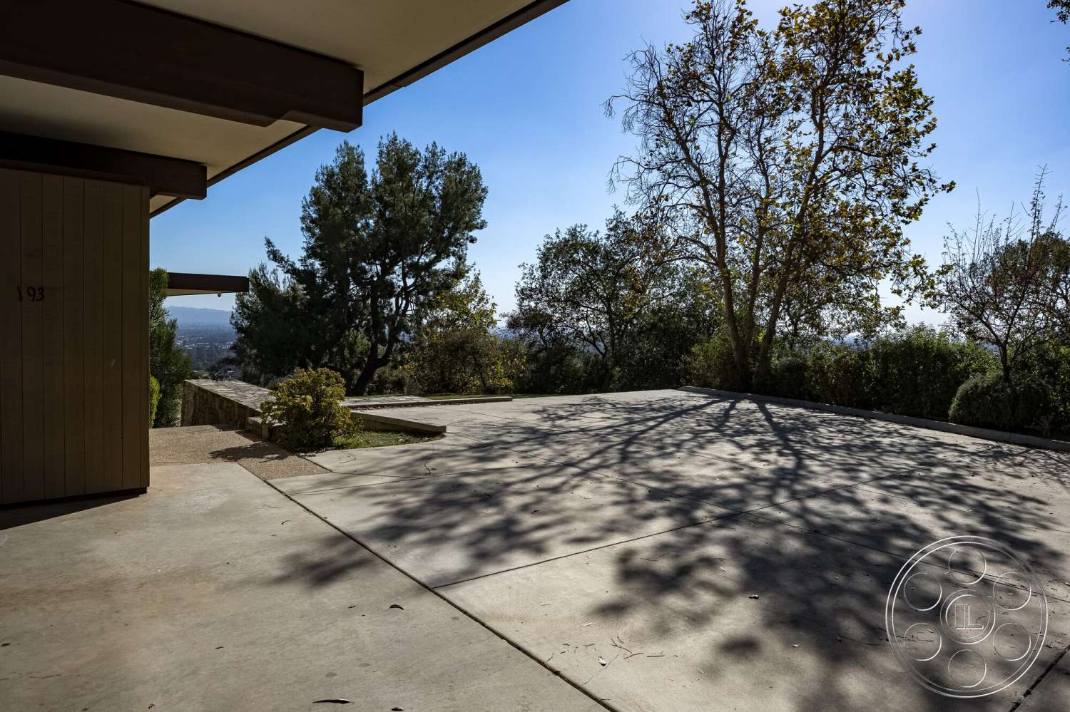 MCM 29 - natural light, exterior, clear blue sky, urban view, landscaped garden area, modern home design, concrete patio floor, stone garden wall, wood beam overhang, view of surrounding trees, flat rectangular patio, southern hillside location