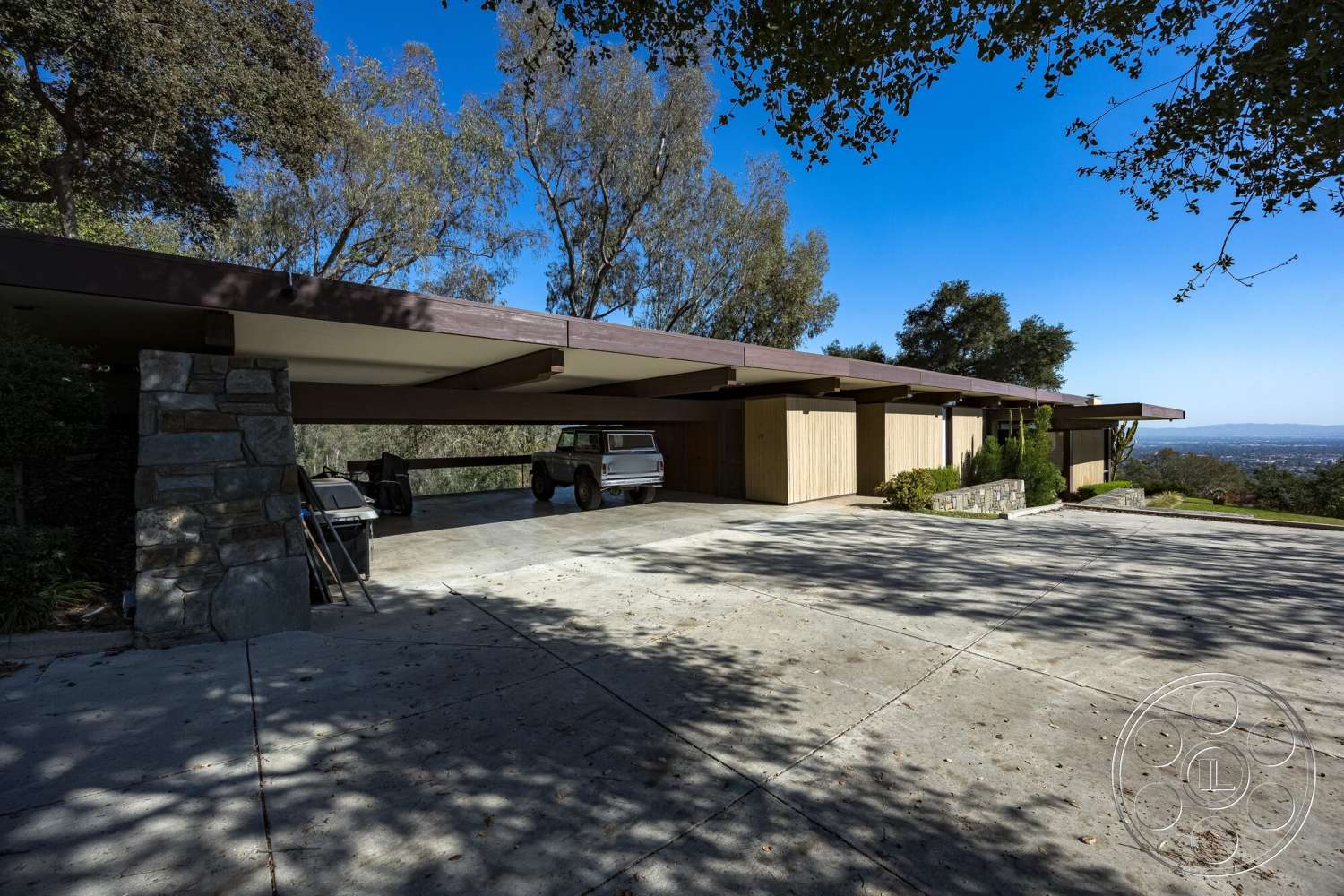 MCM 29 - exterior, floor-to-ceiling windows, modern architectural style, concrete driveway, landscaped garden area, open carport design, stone column supports, sloping roof with overhang, horizontal wooden cladding, distant valley view