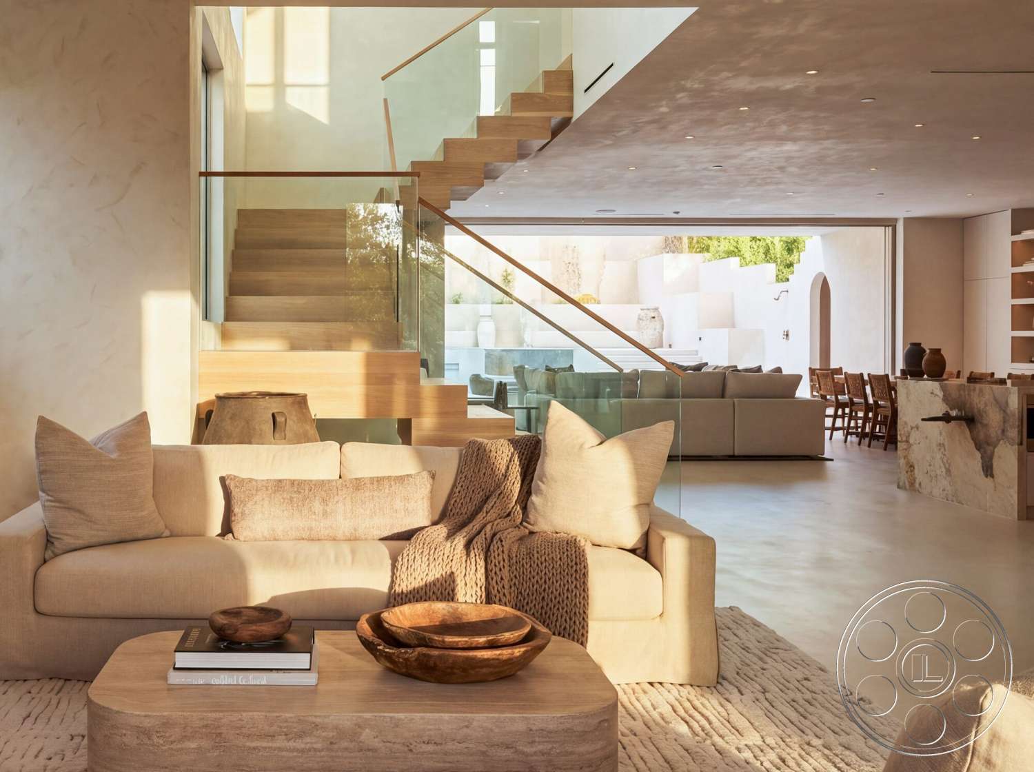 Modern 535 - minimalist decor, neutral color palette, concrete flooring, large floor-to-ceiling windows, beige sectional sofa, textured throw blankets, round wooden coffee table, glass railing staircase, wooden stair treads, wooden dining chairs, natural fiber area rug, exposed plaster walls, modern kitchen island with stone countertop, open plan living area