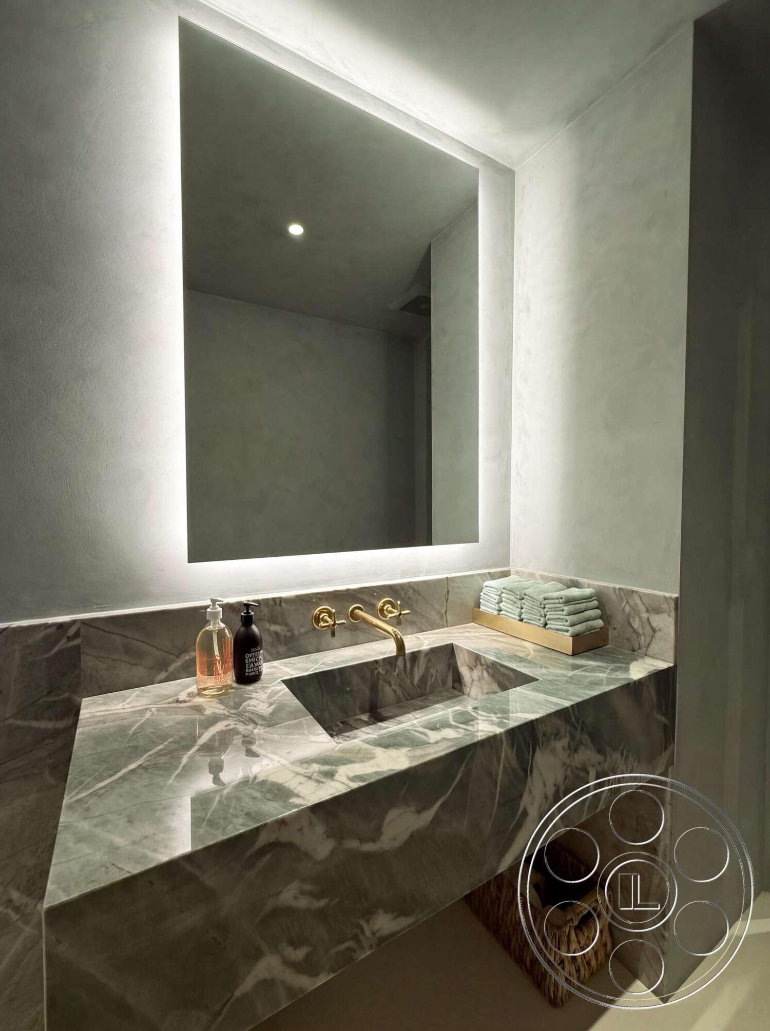 Modern 535 - minimalist design, textured plaster walls, woven storage basket, gold faucet fixtures, marble bathroom countertop, built-in mirror with backlighting, bath towels stacked, rectangular sink cutout