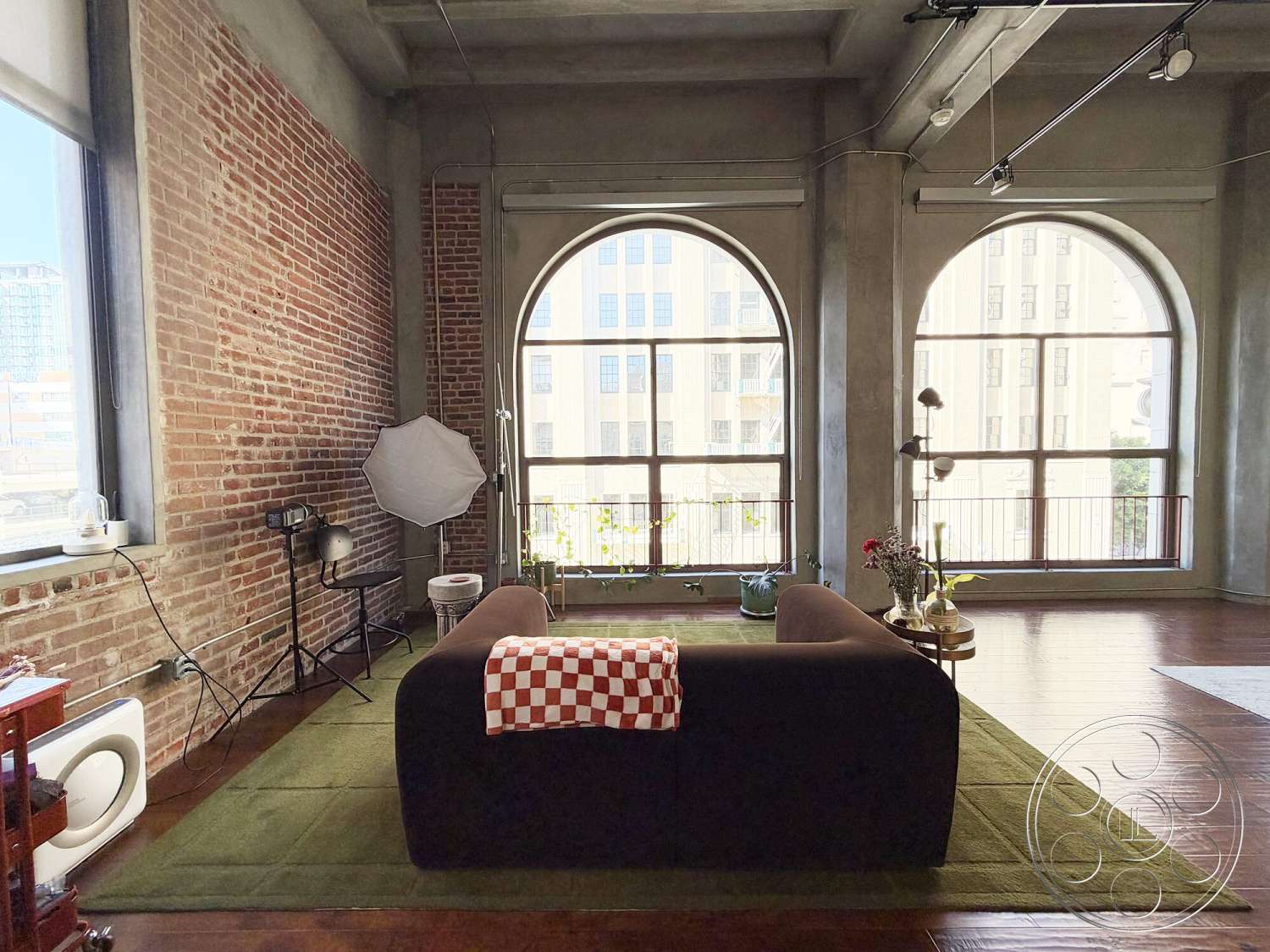 Brockman 310 - hardwood flooring, floor-to-ceiling windows, industrial lighting fixtures, concrete ceiling, arched windows, green area rug, open-concept living area, modern sofa, exposed brick accent wall, urban loft setting