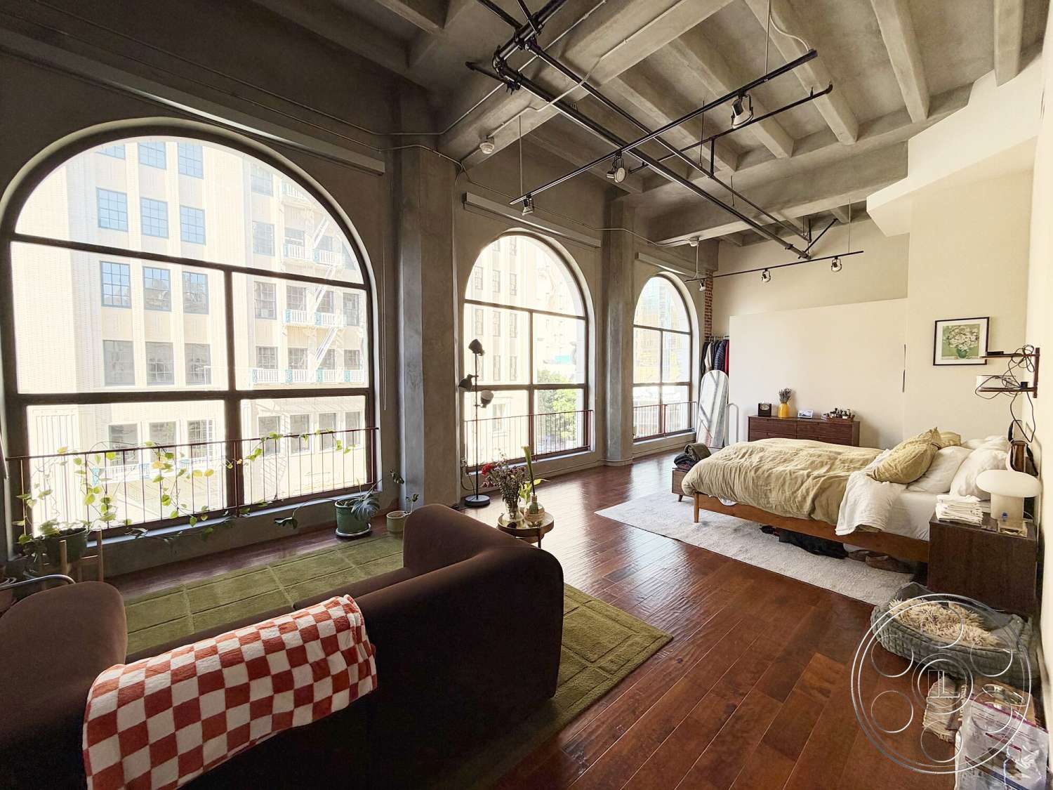 Brockman 310 - textured area rug, floor-to-ceiling windows, arched windows, soft textile bedding, urban apartment setting, exposed concrete ceiling beams, modern bedroom furniture, wooden bedroom floors, plant-filled window ledge