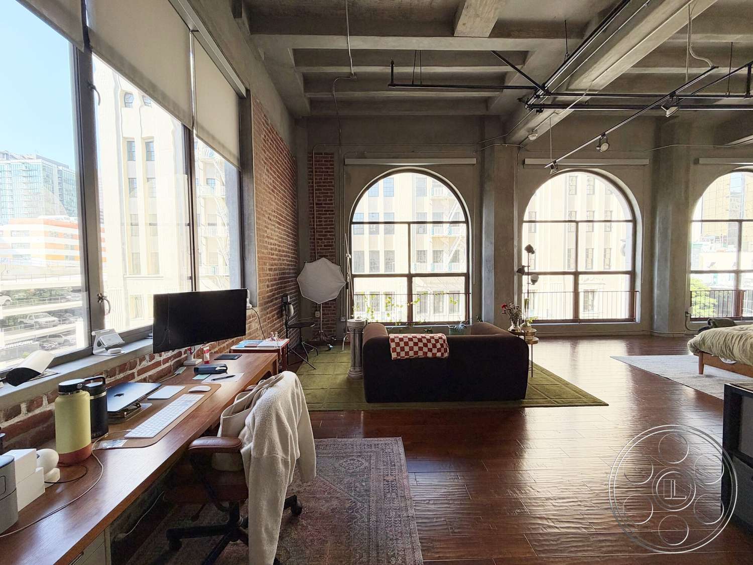 Brockman 310 - floor-to-ceiling windows, open concept living space, hardwood living room floors, modern sofa, decorative indoor plants, exposed brick accent wall, urban city view, industrial style windows, concrete ceiling beams, curved coffee table, wood desk with ergonomic chair, studio apartment layout, oversized armchair
