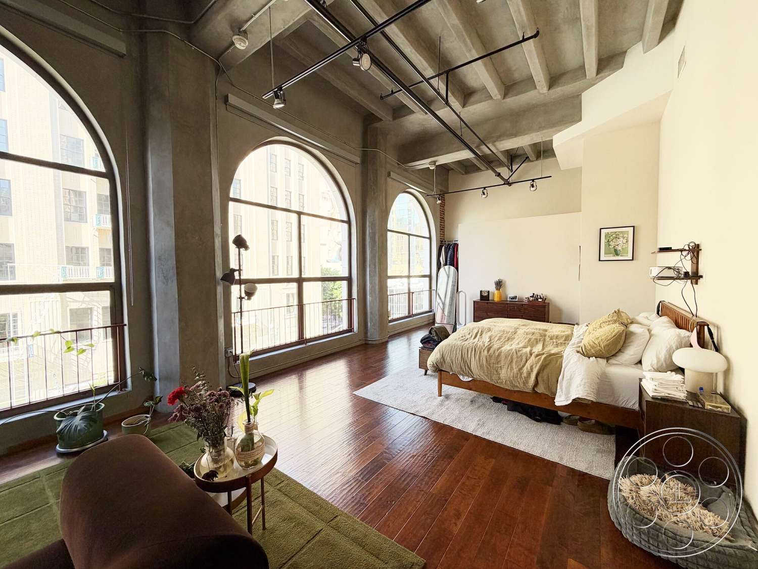 Brockman 310 - floor-to-ceiling windows, soft area rug, textured plaster walls, open living space, exposed concrete ceiling, arched windows, hardwood bedroom floors, minimalist bedroom decor, decorative indoor plants, industrial-style interior, metal track lighting fixtures, king-size bed with patterned bedding, wooden dresser with decorative items