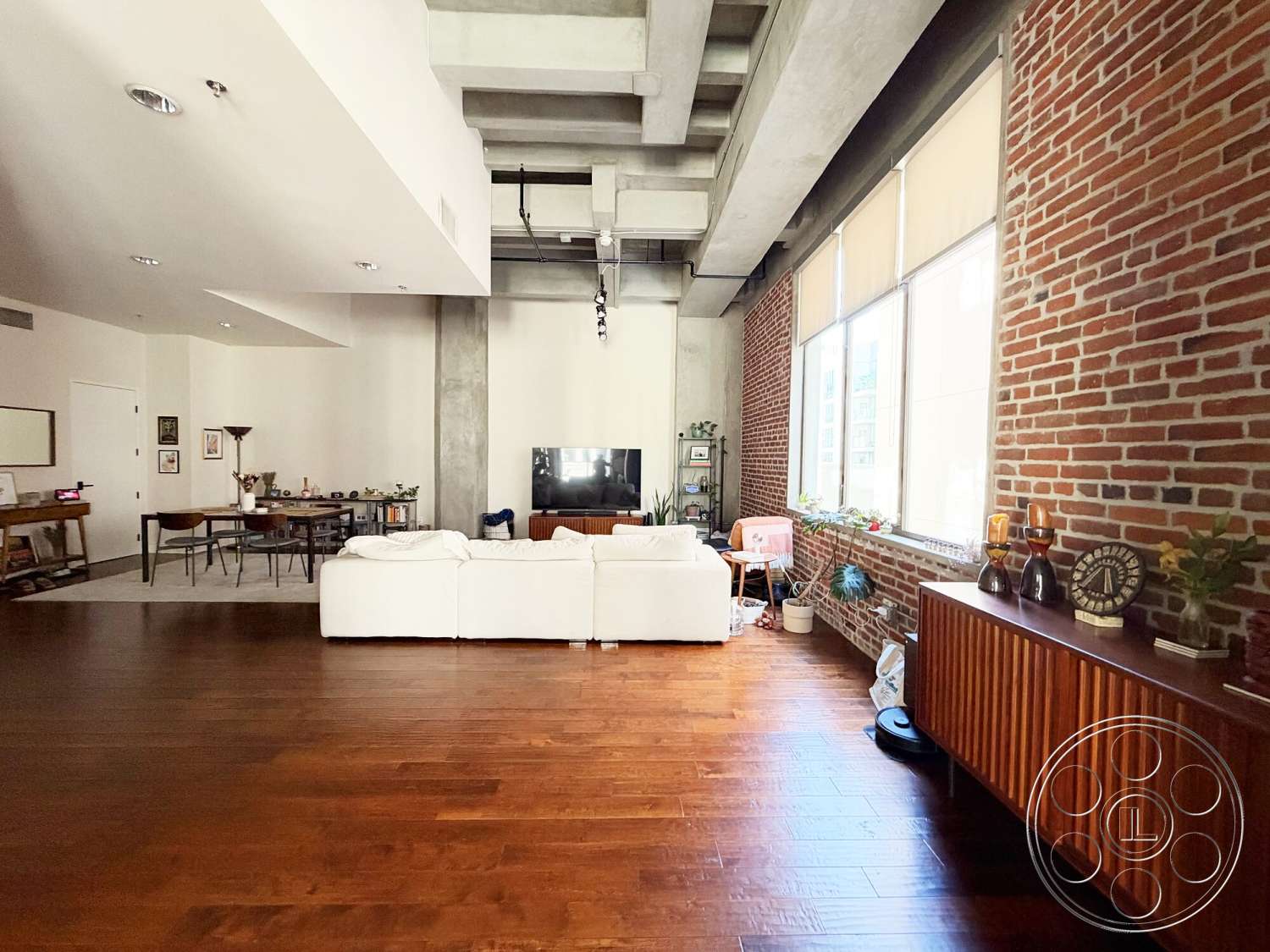 Brockman 310 - large floor-to-ceiling windows, hardwood living room floors, neutral painted walls, exposed brick accent wall, urban loft setting, contemporary home decor, modern white sectional sofa, concrete ceiling with exposed beams, industrial-style table and chairs, wooden storage cabinet with vertical slats