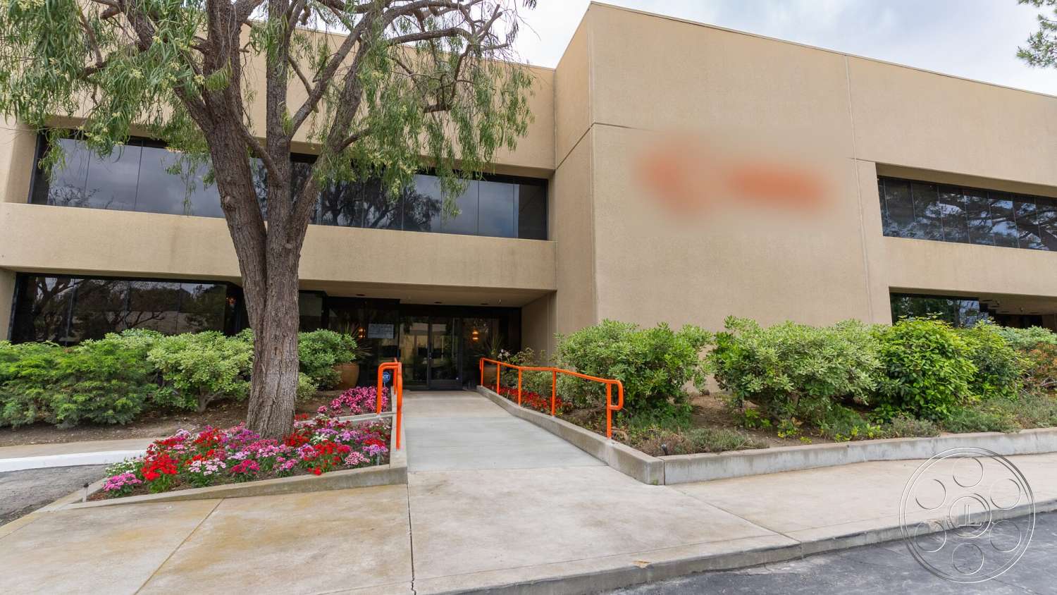 Gym 38 - exterior, large glass windows, metal railing, concrete walkway, landscaped garden area, concrete curb, flower bed with colorful blooms, mature tree, stucco wall facade, orange accent handrails
