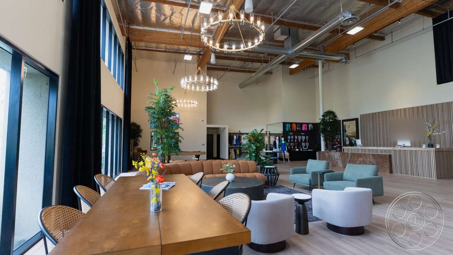 Gym 38 - hardwood flooring, open floor plan, potted greenery, textured wall paneling, large floor-to-ceiling windows, upholstered seating, light wood dining table, exposed wood ceiling beams, contemporary lounge area, reception area with wooden counter, metal ductwork ceiling, modern pendant chandeliers, colorful clothing display section