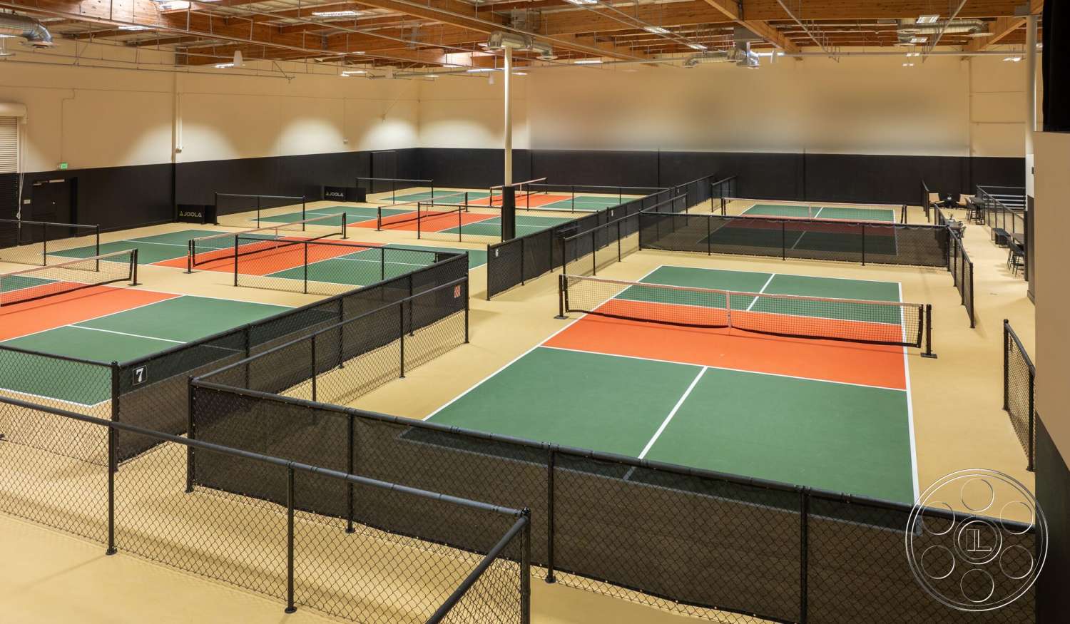 Gym 38 - exposed wood beams, black painted walls, acoustic paneling, LED ceiling lights, green artificial grass flooring, black chain-link fencing, indoor tennis courts, orange game court sections, high vaulted wooden ceiling