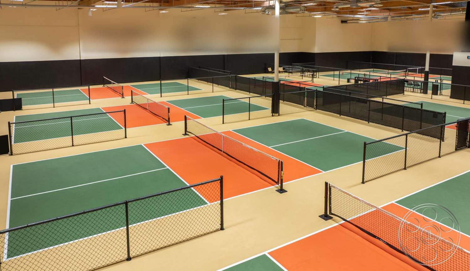 Gym 38 - high ceilings, industrial lighting fixtures, black painted walls, interior sports facility, metal support poles, indoor tennis courts, green synthetic tennis court surface, orange accent court sections, beige polyurethane court flooring, black chain link fences, white court boundary lines, wooden ceiling structure