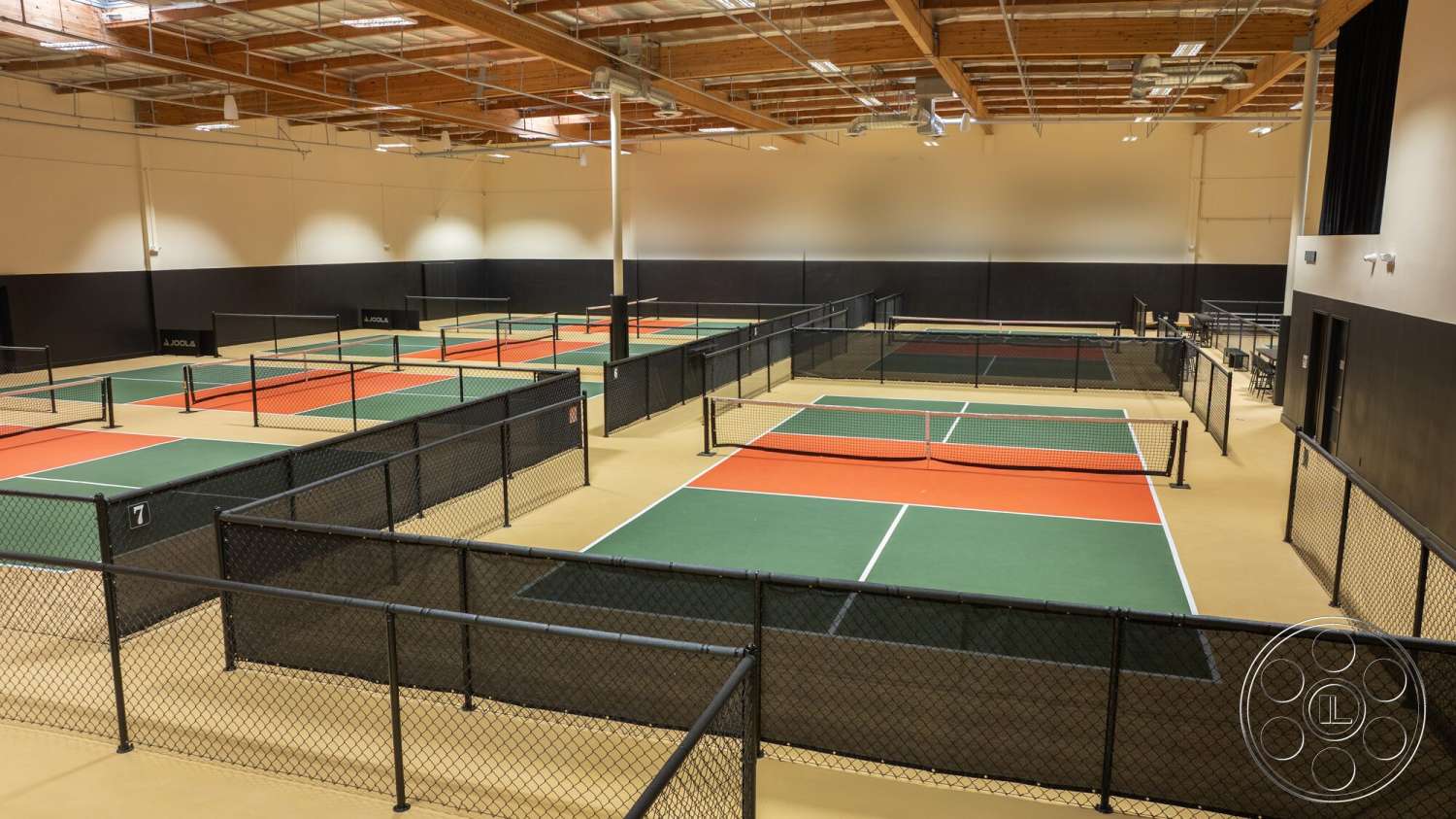 Gym 38 - high ceilings, seating area, industrial lighting fixtures, exposed ceiling structure, wood beam ceiling, gray painted walls, orange court markings, black mesh fencing, green pickleball courts, vinyl sport flooring, multicourt setup
