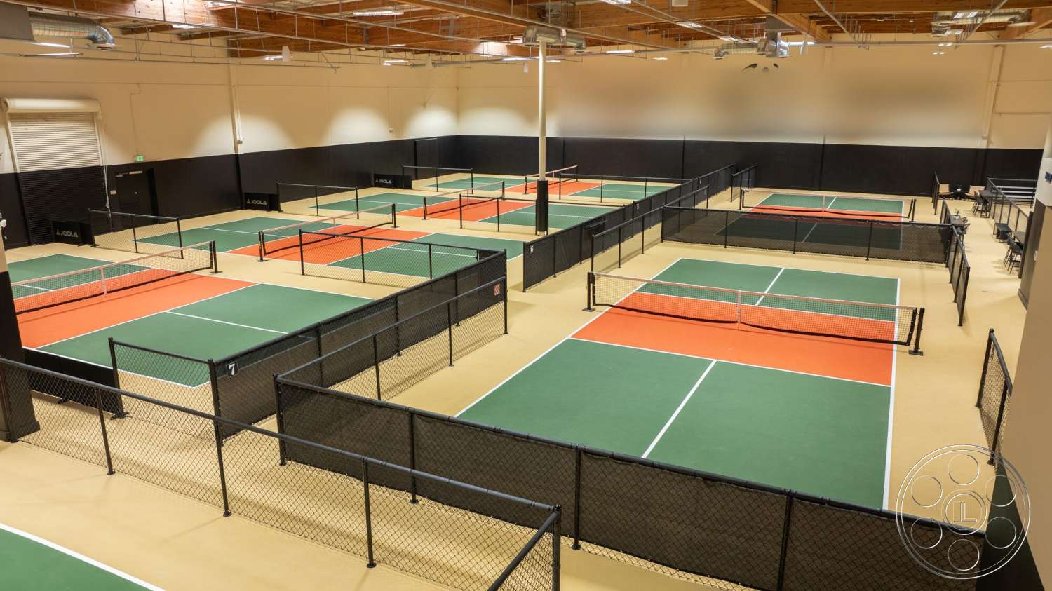 Gym 38 - exposed wooden ceiling beams, black wall paneling, rubberized sports flooring, green pickleball court surfaces, orange pickleball court markings, black vinyl-coated chain-link fencing, high ceiling athletic space, multiple pickleball courts layout, commercial sports facility