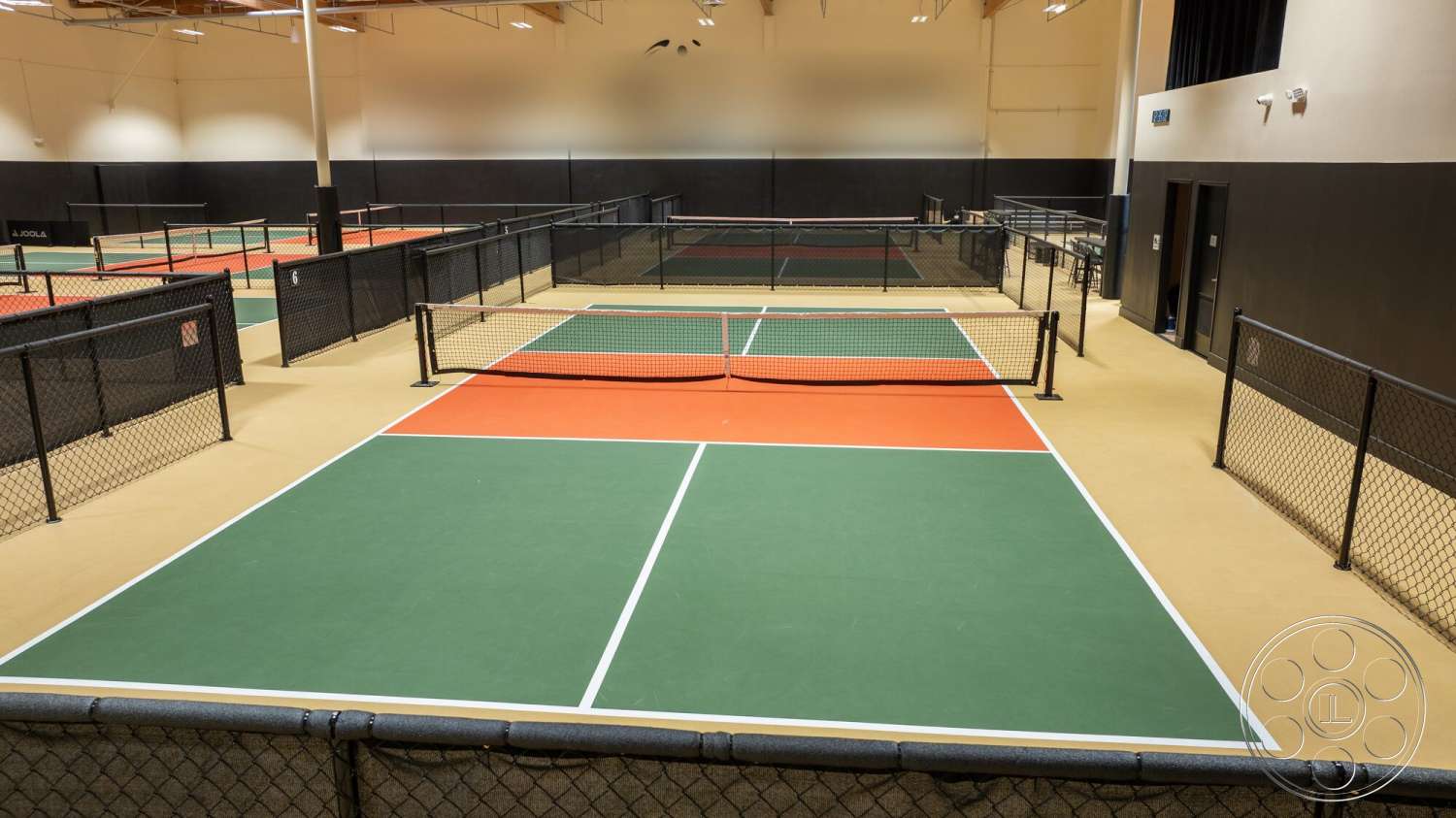 Gym 38 - high ceilings, hardwood flooring, exposed beams, green tennis court surface, LED lighting fixtures, white-painted walls, sports facility interior, grey wall panels, black chain-link fencing, indoor tennis courts, orange court sections, court nets