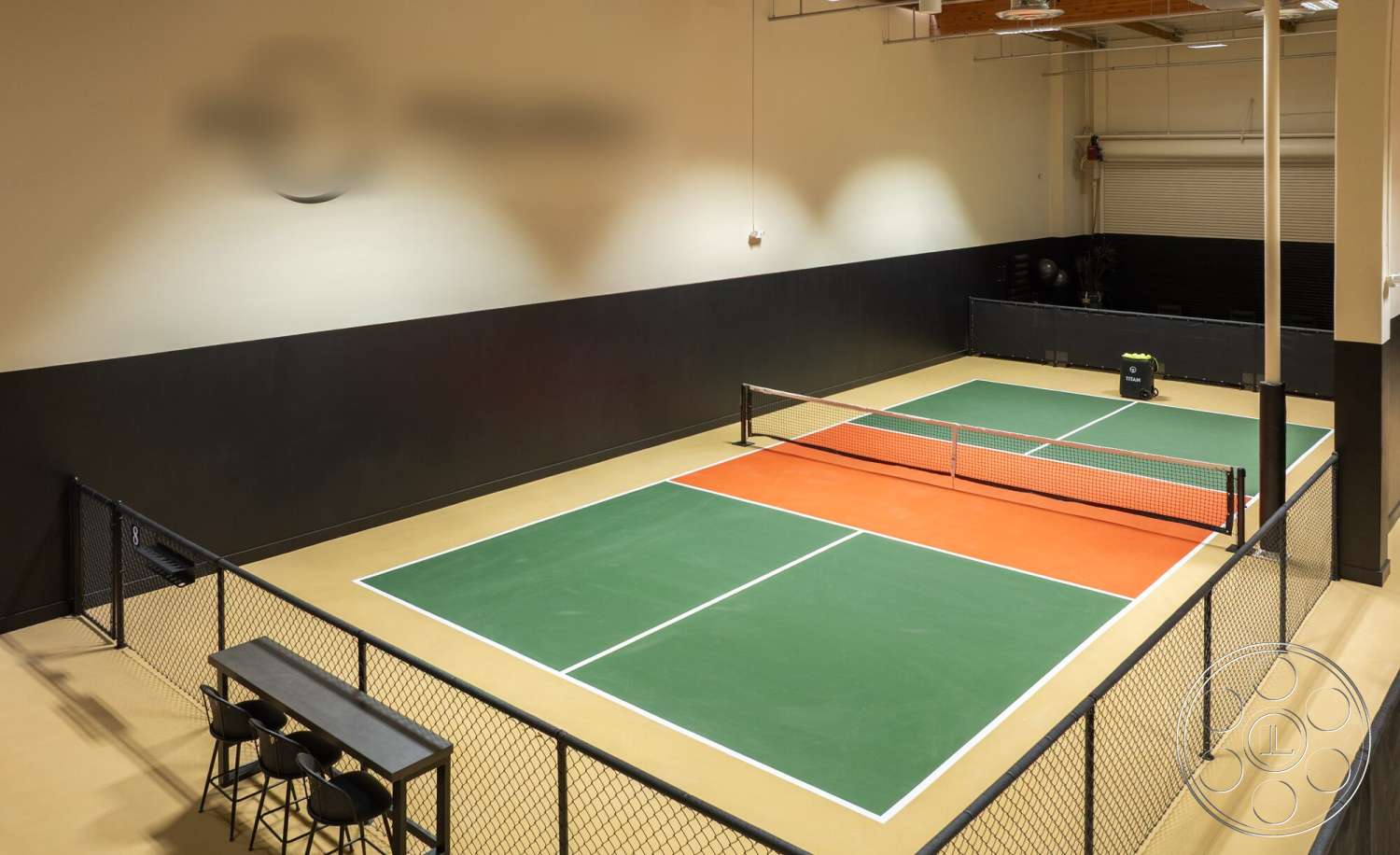 Gym 38 - exposed wood ceiling beams, minimalist seating area, studio lighting fixtures, interior tennis court flooring, green sport court surface, orange court accent area, black vinyl walls, metal chain link fencing