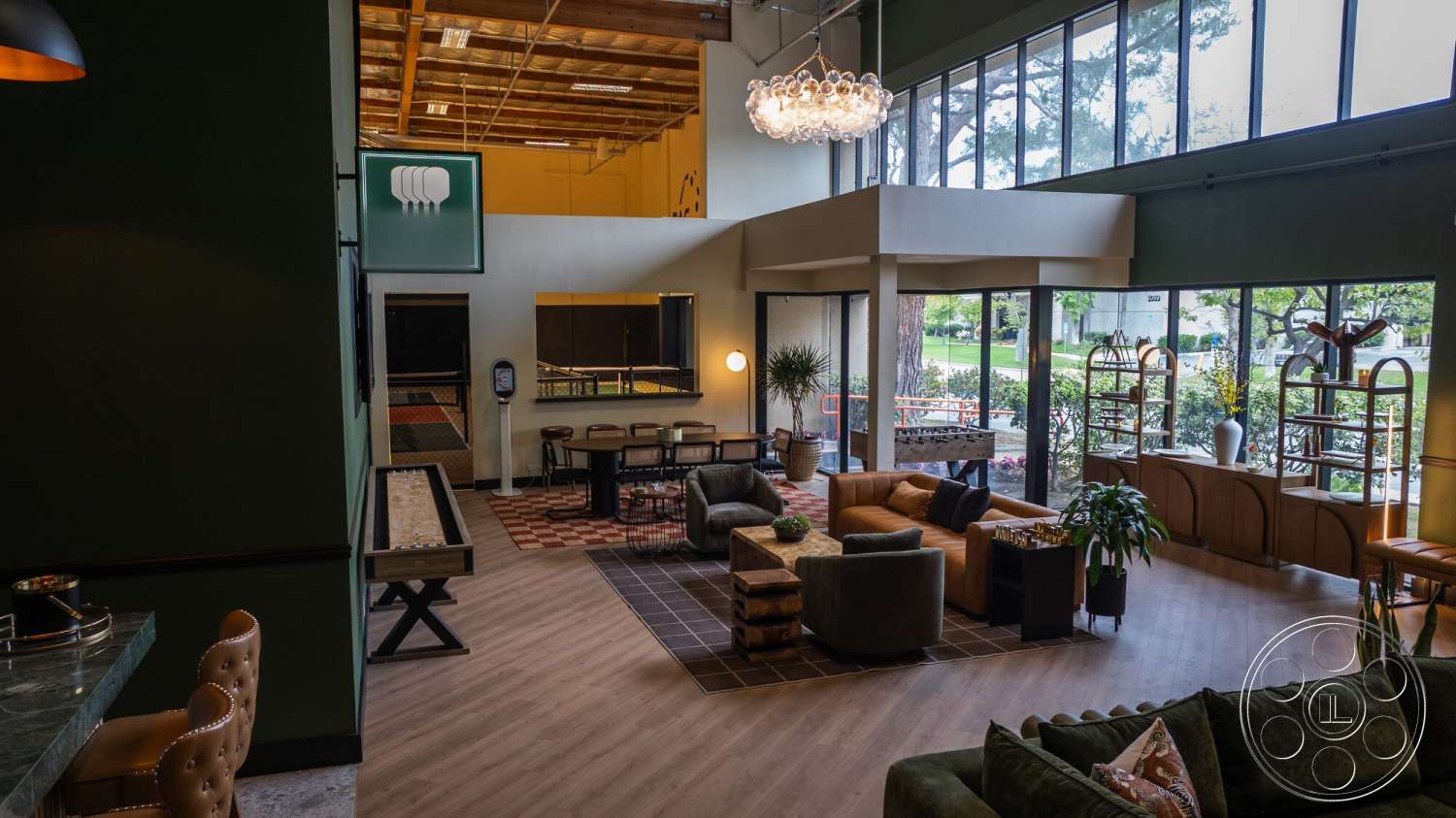 Gym 38 - textured area rug, light hardwood flooring, green accent wall, large floor-to-ceiling windows, rectangular coffee table, plush upholstered sofa, inviting lounge area, high ceiling with exposed wood beams, modern pendant chandelier, round velvet accent chairs, metal and wood shelving units, cushioned bar stool, art deco-inspired decor