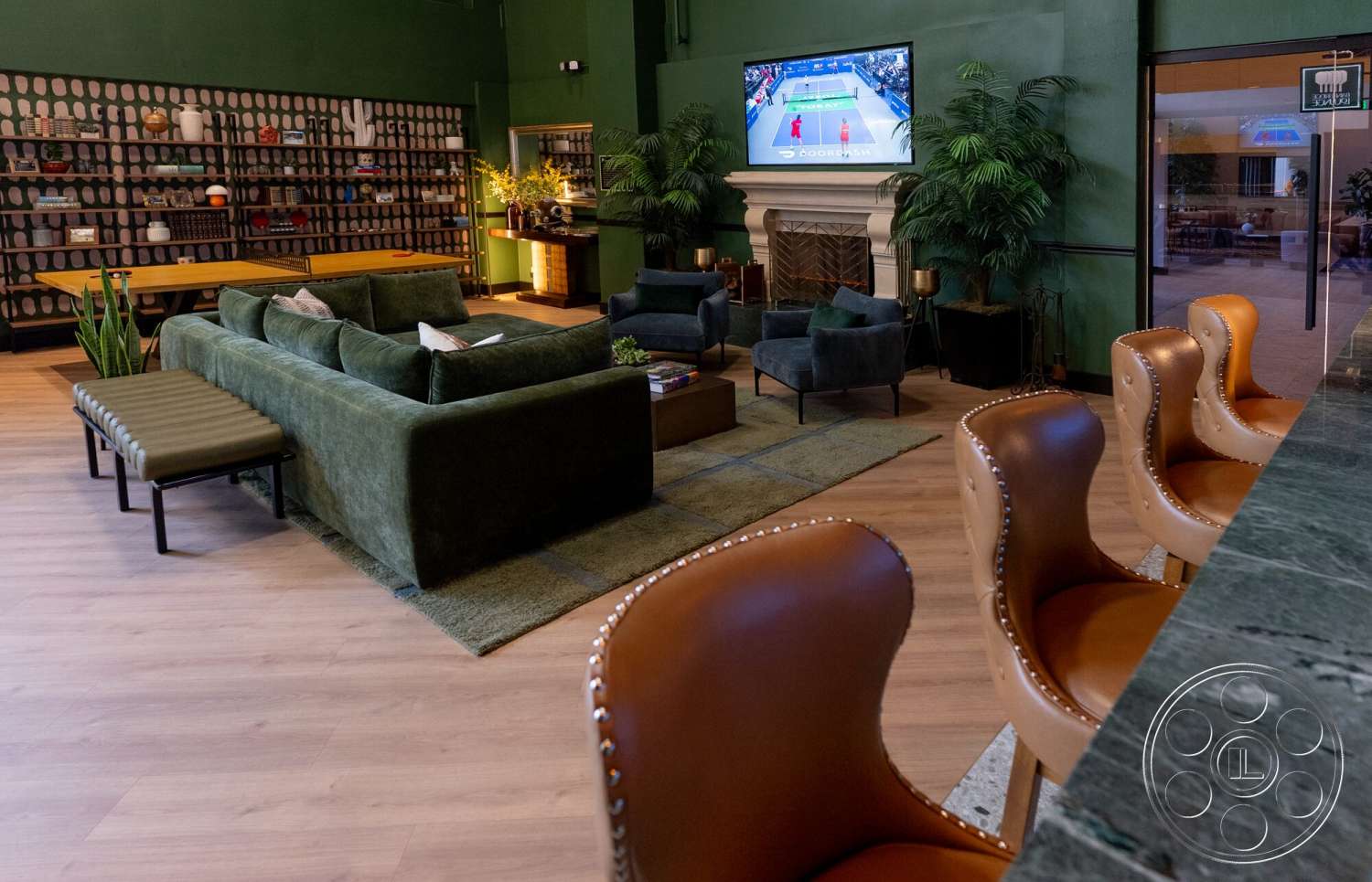 Gym 38 - hardwood flooring, textured area rug, wooden dining table, marble countertop, large indoor plants, exposed shelving units, green painted walls, plush green sectional sofa, leather bar stools, modern fireplace with white mantel, fabric accent chairs, floor-length windows