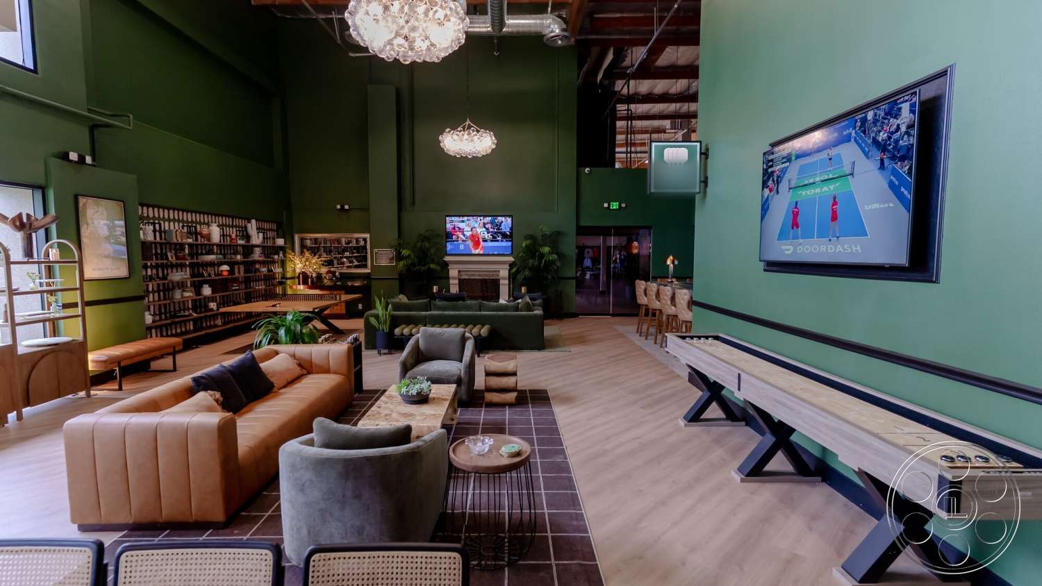 Gym 38 - hardwood flooring, decorative wall art, exposed ceiling beams, lush indoor plants, chandelier lighting fixture, green painted walls, open shelving unit with books, tan leather sectional sofa, modern wood coffee table, light gray upholstered armchair, black framed television, linear wooden bench, black trim around windows
