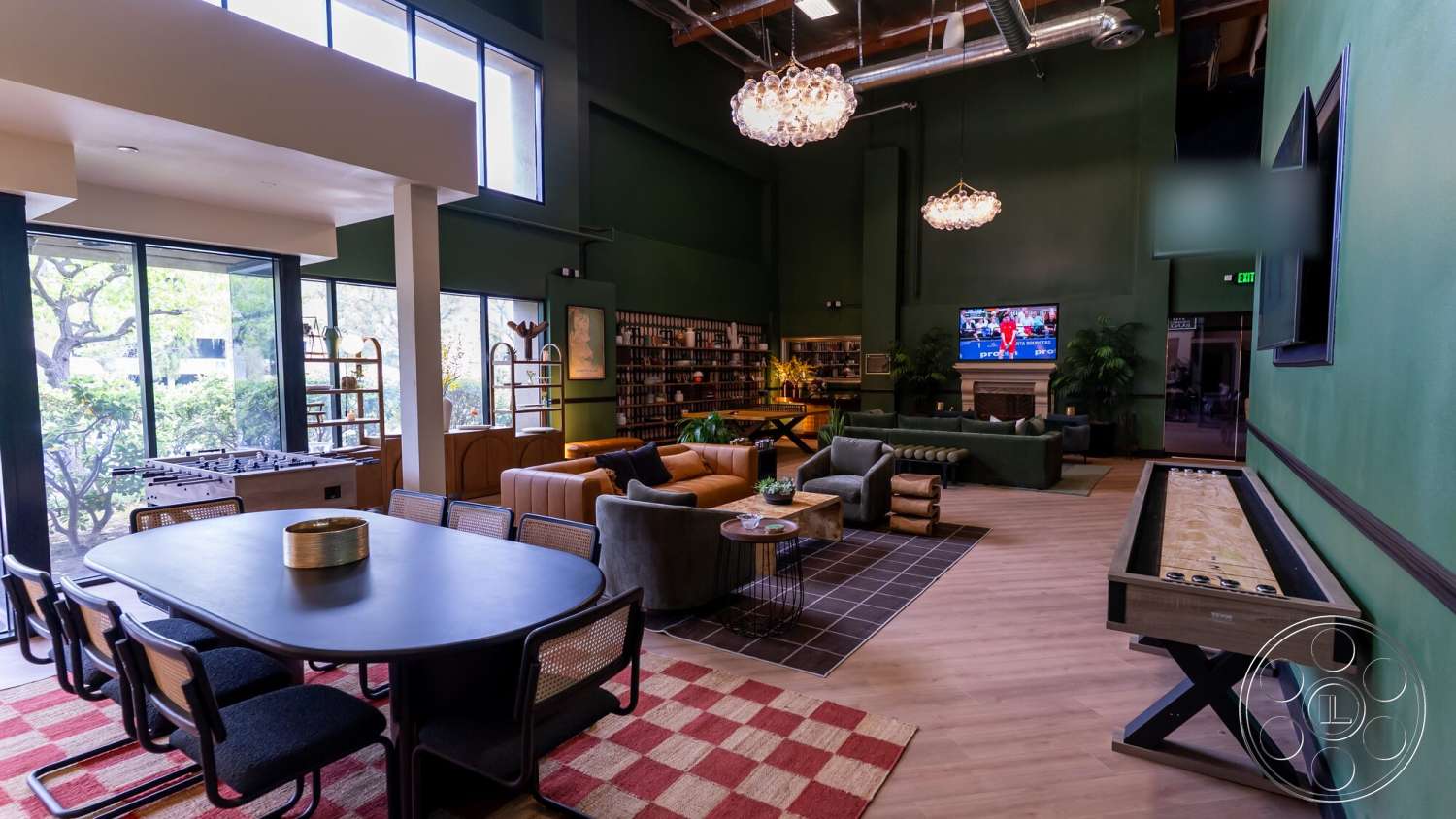 Gym 38 - plush sectional sofa, greenery visible through windows, open concept living space, large floor-to-ceiling windows, contemporary coffee table, exposed wood ceiling beams, hardwood flooring in living room, industrial-style lighting fixtures, vaulted ceiling in living area, dark green painted walls, textured area rug with geometric pattern, cozy sitting area with accent pillows, minimalist dining table with black surface, game table for leisure activities, built-in shelving for storage